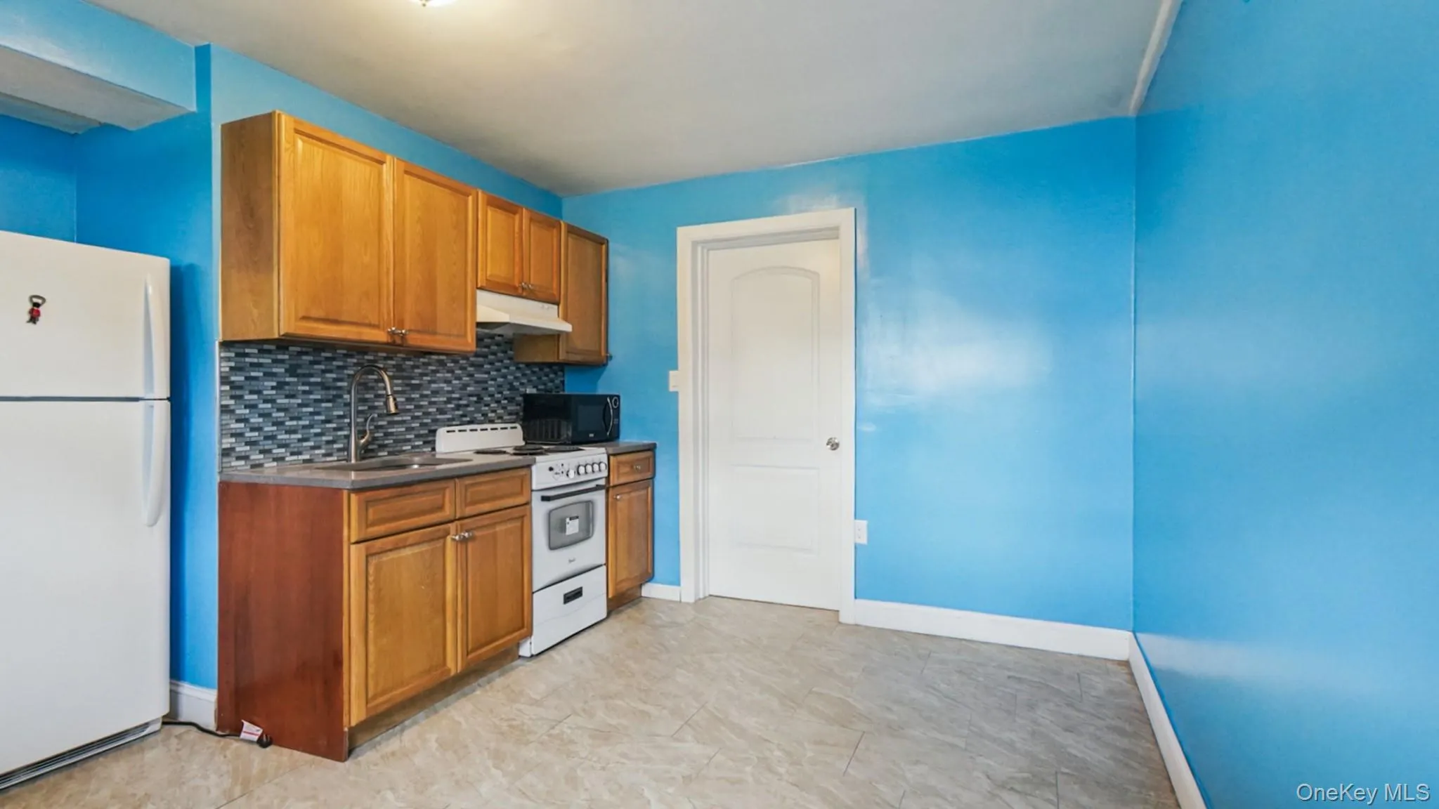 Kitchenette featuring wood-finish cabinetry, a patterned tile backsplash, stainless steel sink, and white appliances Kitchenette featuring wood-finish cabinetry, a patterned tile backsplash, stainless steel sink, and white appliances