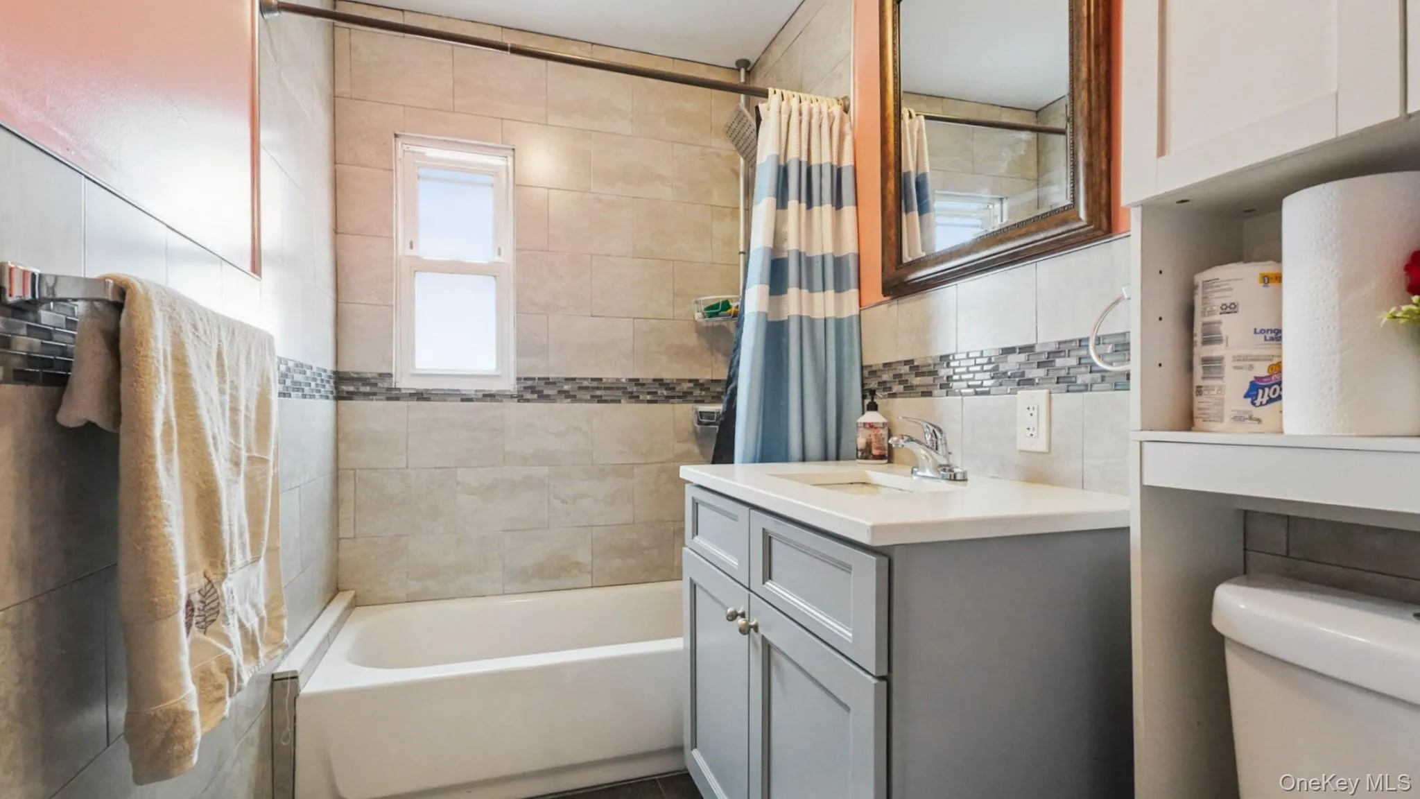 Bathroom featuring a light gray vanity with a white countertop, a white bathtub, and floor-to-ceiling tiled walls with a mosaic accent band Bathroom featuring a light gray vanity with a white countertop, a white bathtub, and floor-to-ceiling tiled walls with a mosaic accent band