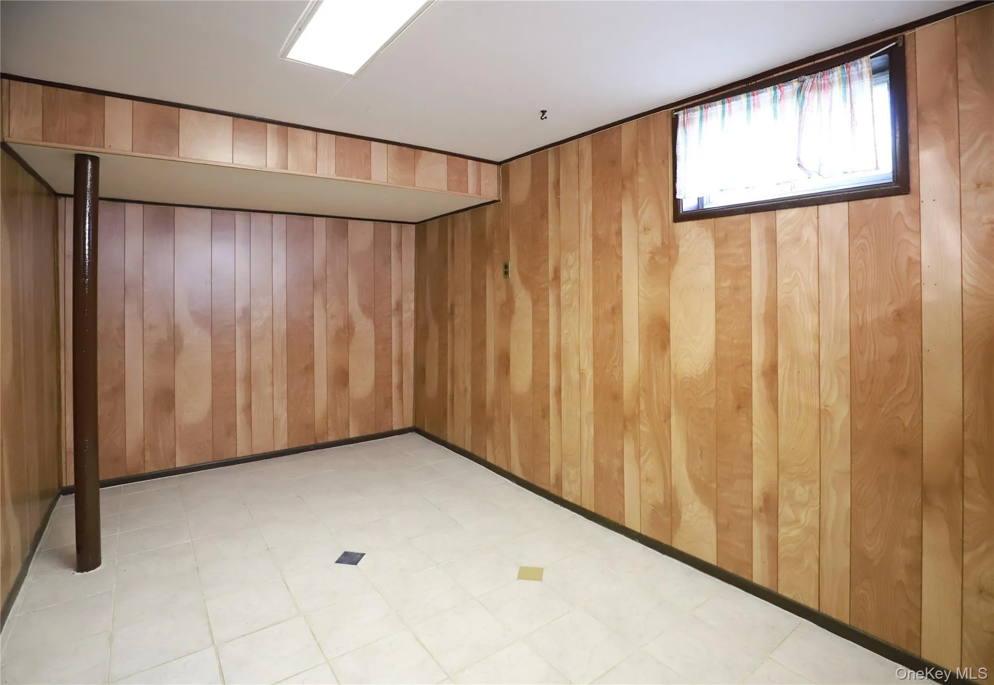 Wood-paneled room featuring light-toned ceramic tile flooring, a rectangular window, and a fluorescent ceiling light fixture Wood-paneled room featuring light-toned ceramic tile flooring, a rectangular window, and a fluorescent ceiling light fixture
