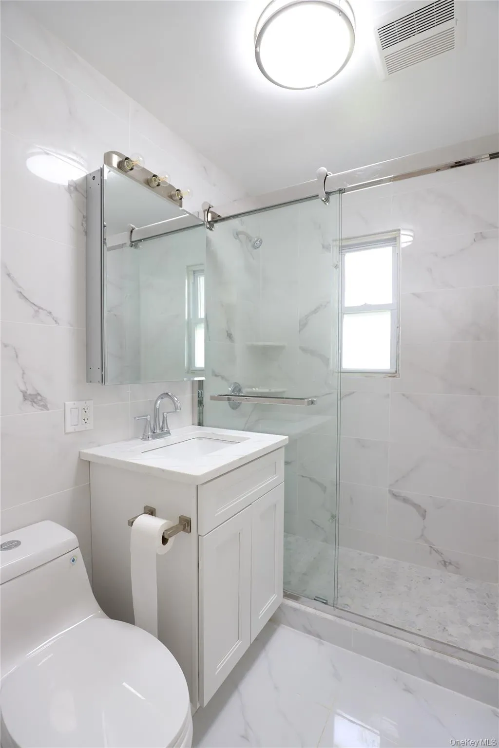 Bathroom featuring a glass-enclosed shower with sliding door, white vanity with integrated sink, chrome fixtures, and a wall-mounted medicine cabinet with integrated lighting Bathroom featuring a glass-enclosed shower with sliding door, white vanity with integrated sink, chrome fixtures, and a wall-mounted medicine cabinet with integrated lighting