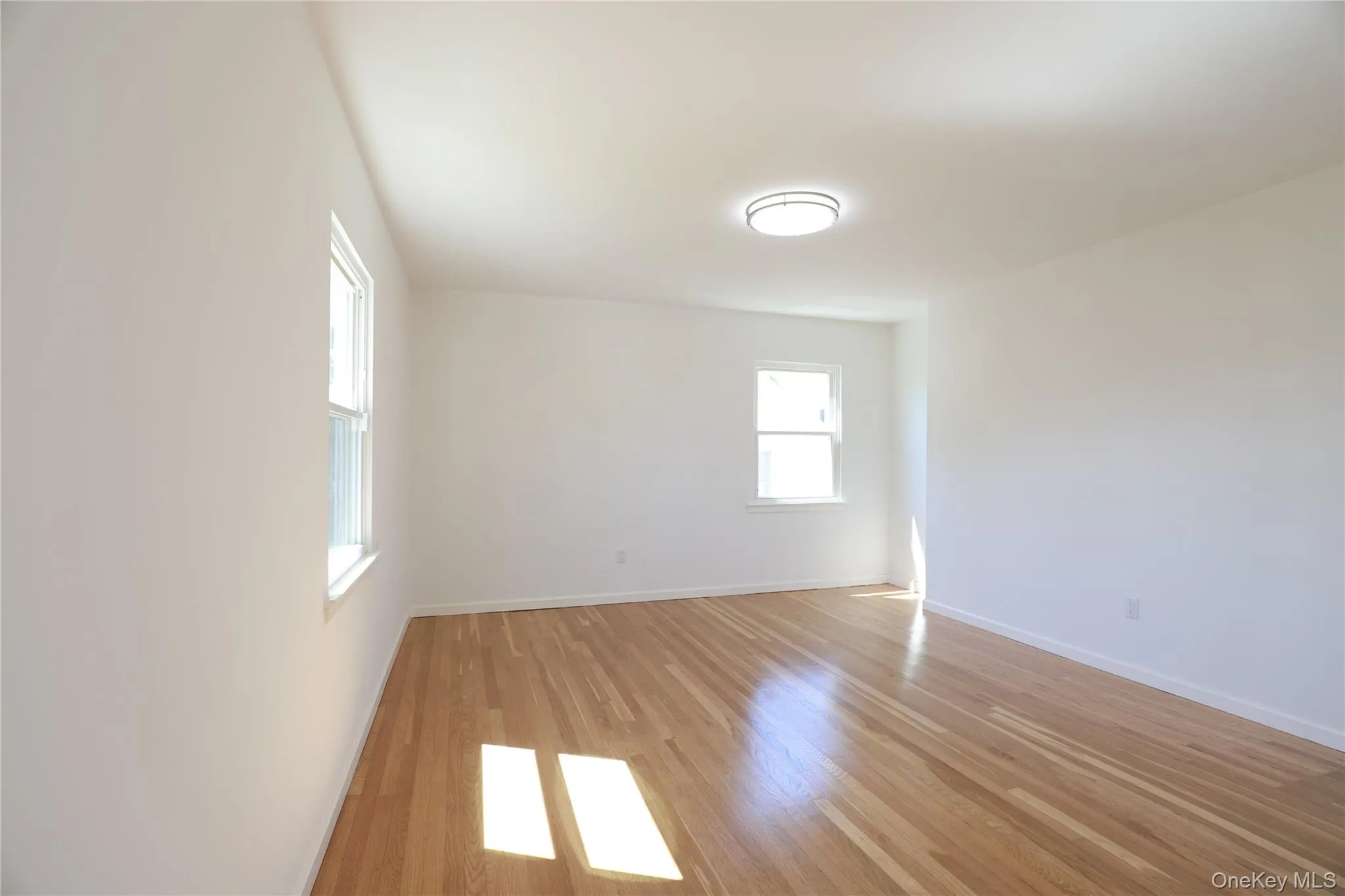 Bright room featuring polished wood-finish flooring, white walls, two windows, a ceiling-mounted light fixture, and a bi-fold door Bright room featuring polished wood-finish flooring, white walls, two windows, a ceiling-mounted light fixture, and a bi-fold door