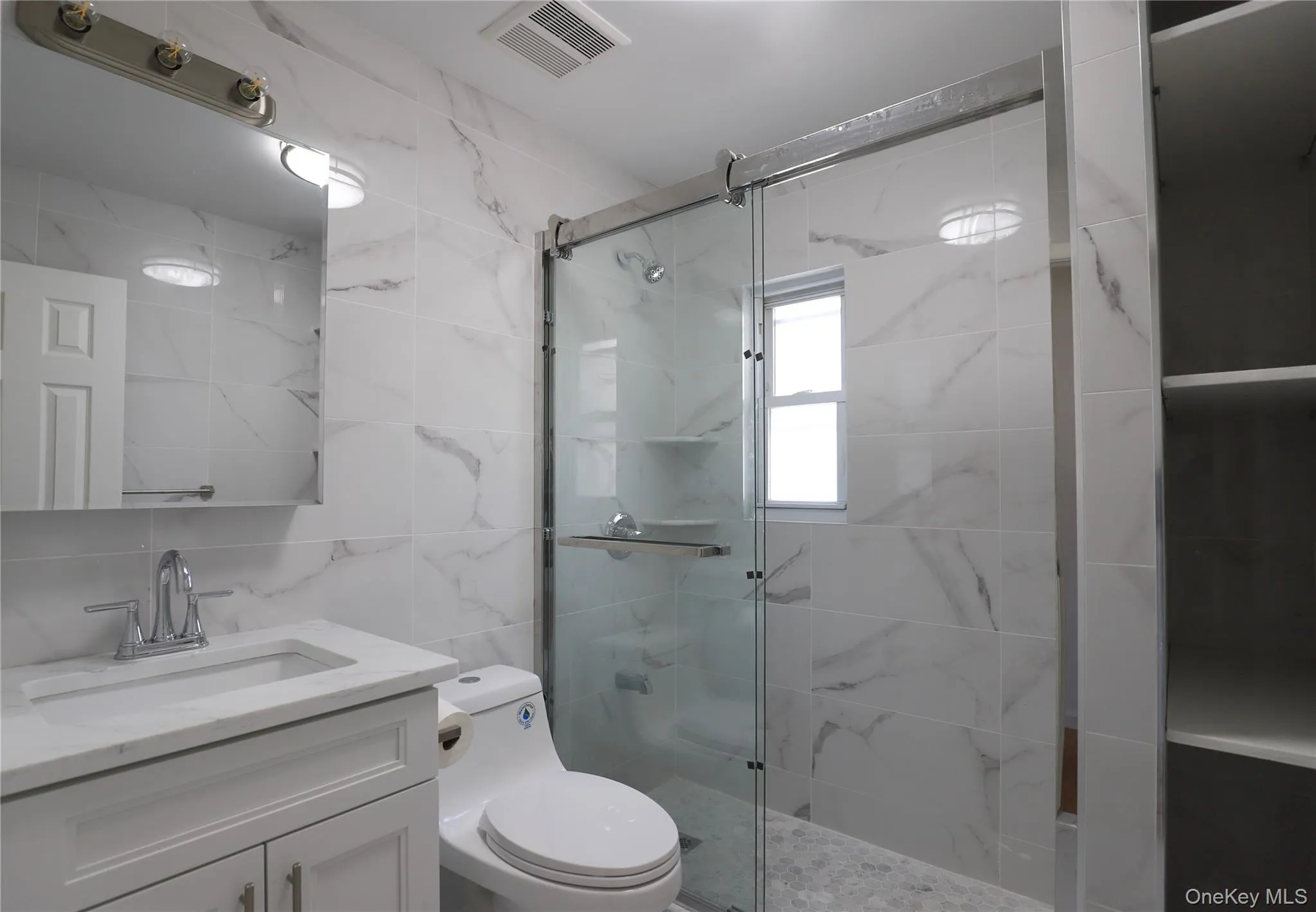 Full bathroom featuring a glass-enclosed shower with pebble-finish flooring, a contemporary white vanity with an integrated sink, polished chrome fixtures, and white tile walls with subtle veining Full bathroom featuring a glass-enclosed shower with pebble-finish flooring, a contemporary white vanity with an integrated sink, polished chrome fixtures, and white tile walls with subtle veining
