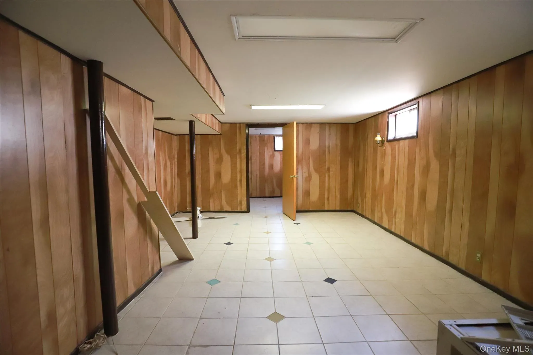 Spacious room featuring wood-finish wall paneling, white ceiling, and light-toned square tile flooring with accent tiles Spacious room featuring wood-finish wall paneling, white ceiling, and light-toned square tile flooring with accent tiles