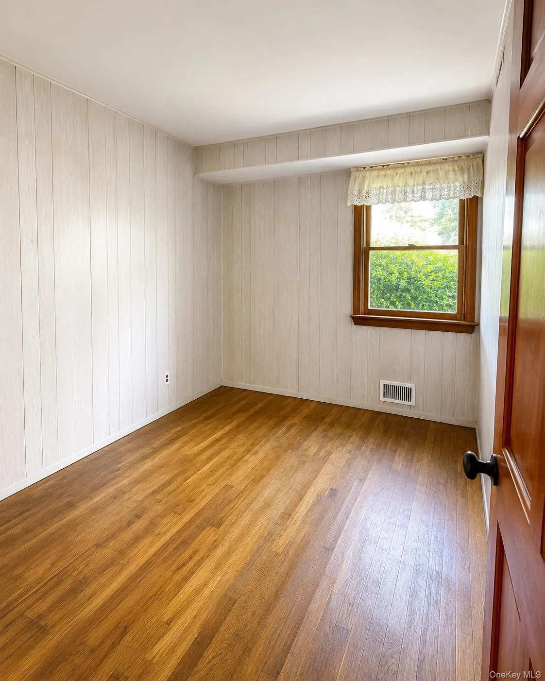 Bedroomfeaturing wood-finish flooring, white vertical paneled walls, and a single window with wood trim Bedroomfeaturing wood-finish flooring, white vertical paneled walls, and a single window with wood trim