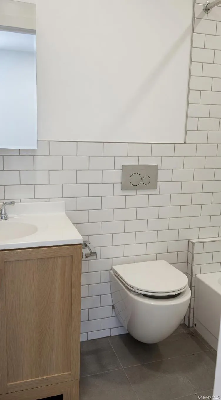 Bathroom featuring subway tile walls, a wall-mounted toilet with a concealed cistern, a light wood-finish vanity with an integrated sink, a mirrored medicine cabinet, and dark gray floor tiles Bathroom featuring subway tile walls, a wall-mounted toilet with a concealed cistern, a light wood-finish vanity with an integrated sink, a mirrored medicine cabinet, and dark gray floor tiles
