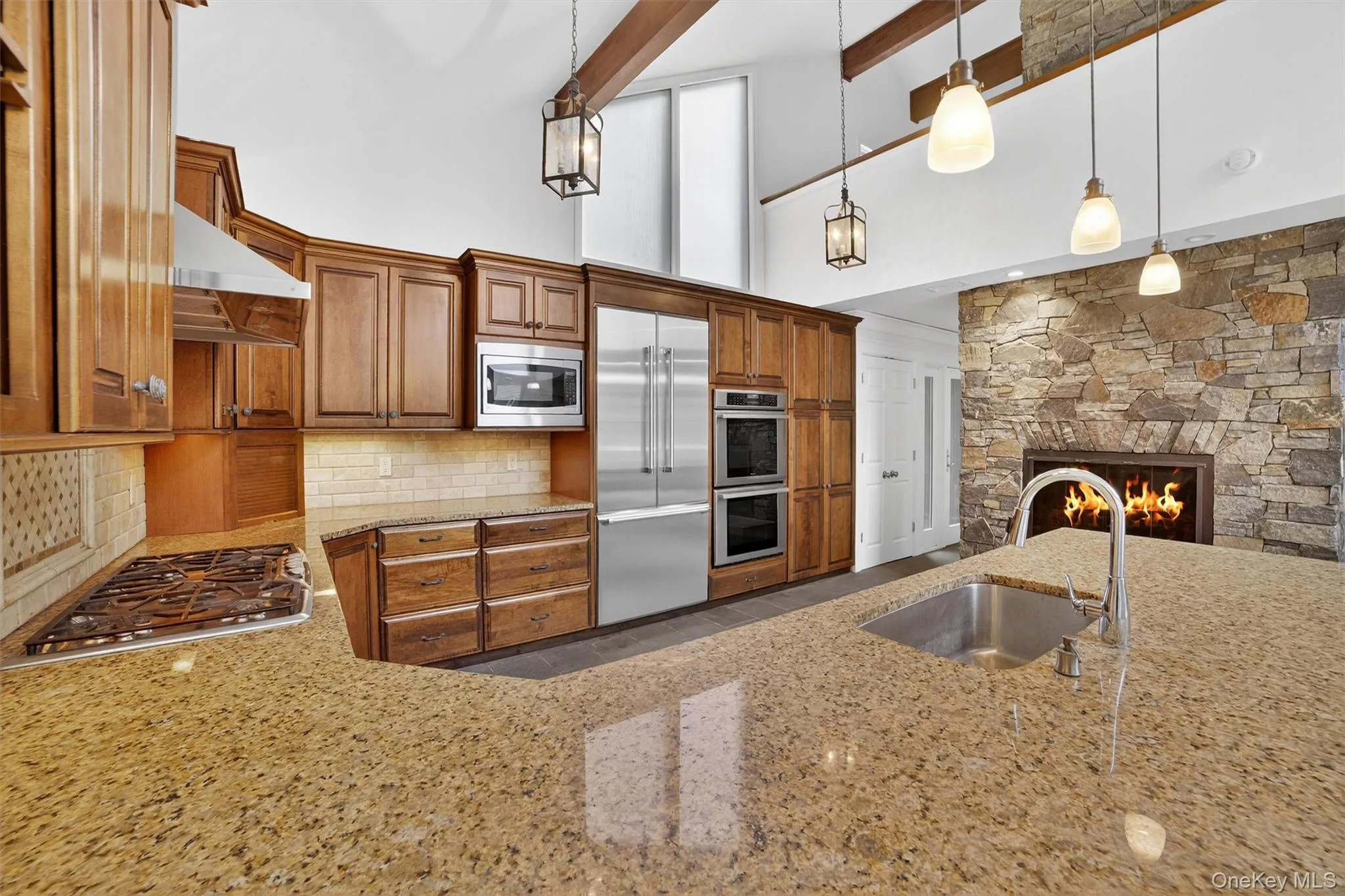 Kitchen featuring built in appliances, light stone countertops, backsplash, under cabinet range hood, and brown cabinets Kitchen featuring built in appliances, light stone countertops, backsplash, under cabinet range hood, and brown cabinets