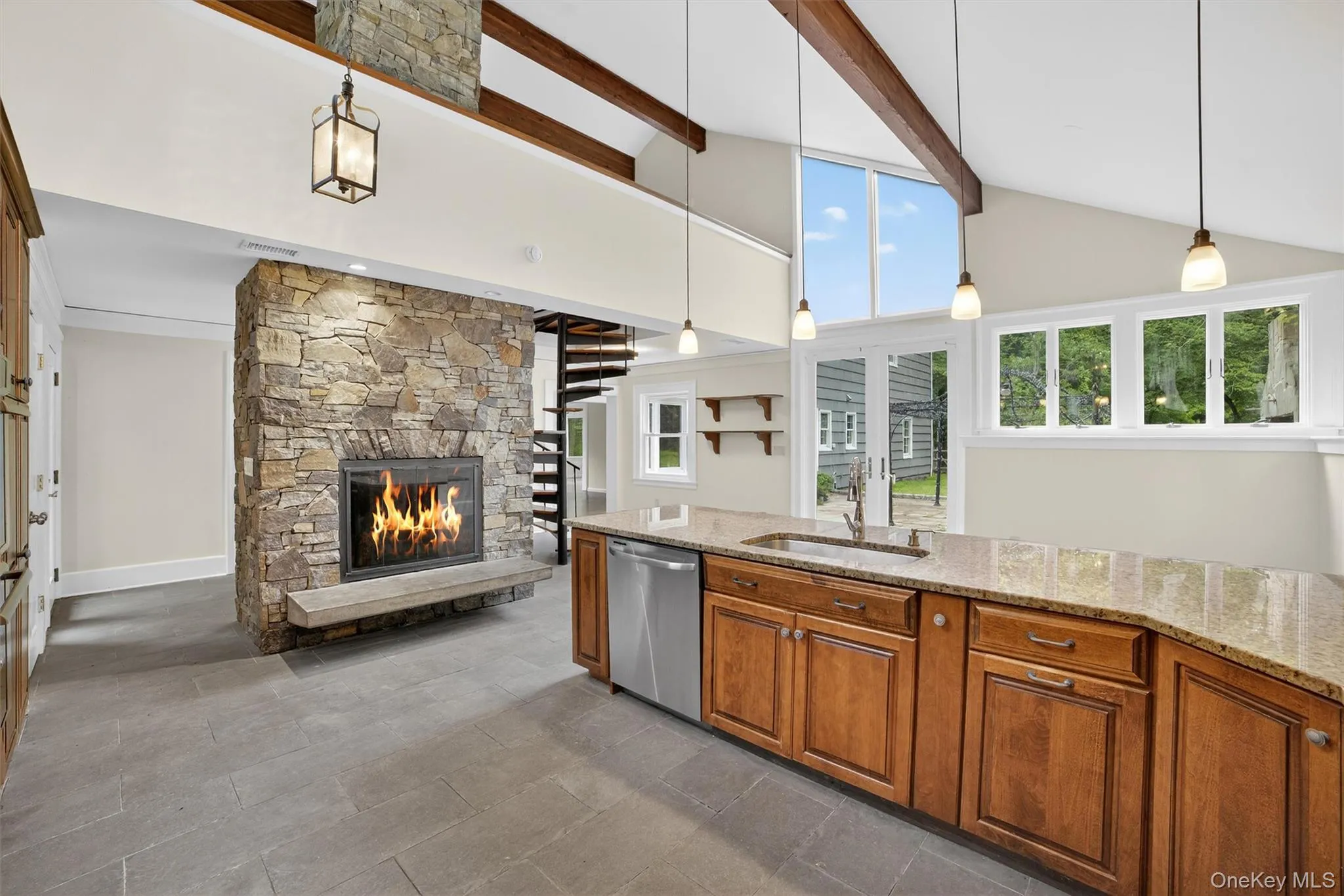 Kitchen featuring stainless steel dishwasher, a stone fireplace, brown cabinetry, beam ceiling, and high vaulted ceiling Kitchen featuring stainless steel dishwasher, a stone fireplace, brown cabinetry, beam ceiling, and high vaulted ceiling