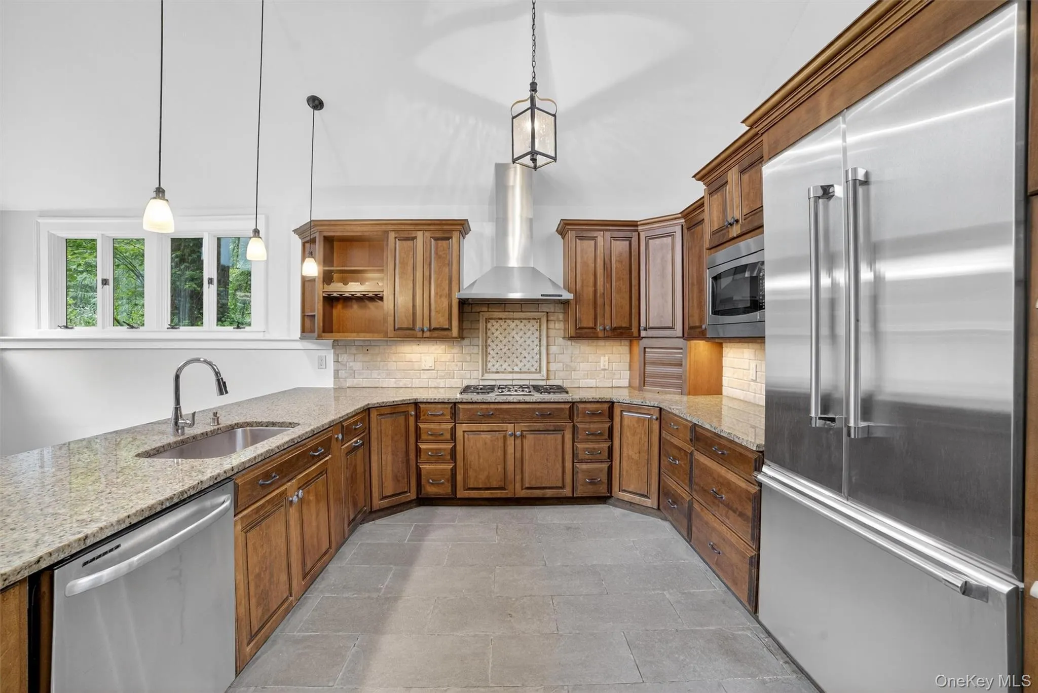 Kitchen with appliances with stainless steel finishes, wall chimney exhaust hood, brown cabinetry, and open shelves Kitchen with appliances with stainless steel finishes, wall chimney exhaust hood, brown cabinetry, and open shelves