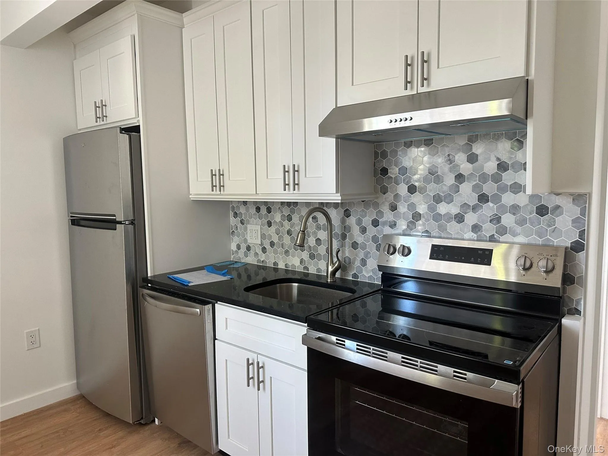 Compact kitchen featuring white shaker cabinetry, stainless steel appliances, black countertop, and a gray hexagonal tile backsplash Compact kitchen featuring white shaker cabinetry, stainless steel appliances, black countertop, and a gray hexagonal tile backsplash