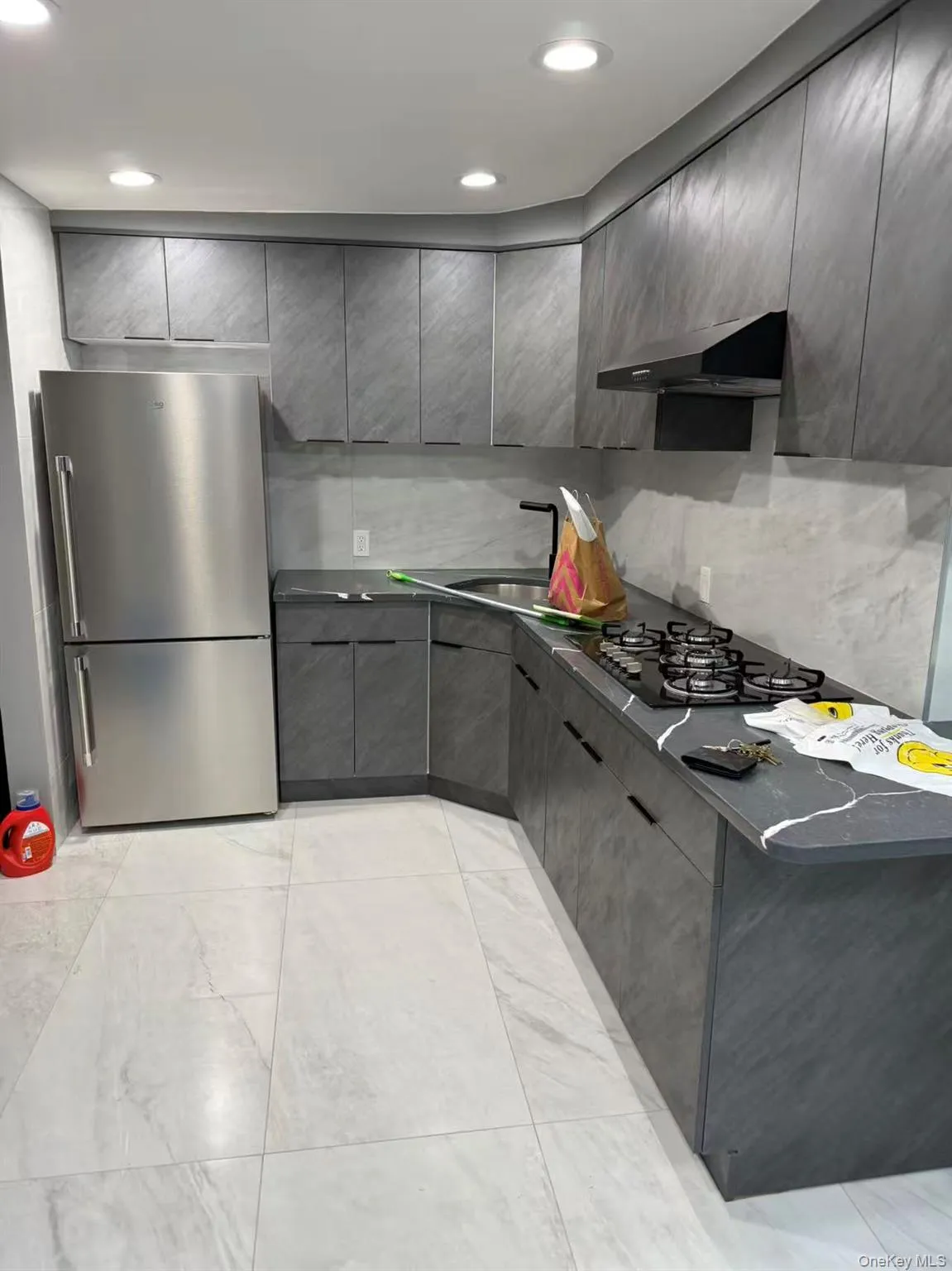 Kitchen with dark countertops, gray cabinetry, freestanding refrigerator, tasteful backsplash, and under cabinet range hood Kitchen with dark countertops, gray cabinetry, freestanding refrigerator, tasteful backsplash, and under cabinet range hood