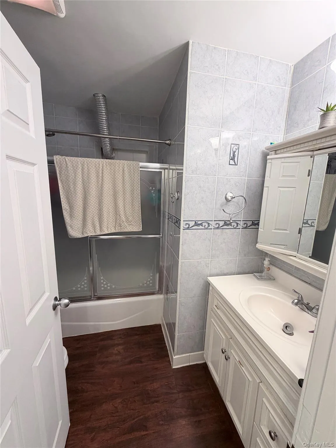 Bathroom featuring white wood-finish vanity with an integrated sink, white tiled walls with decorative accents, wood-finish flooring, and a bathtub with sliding glass doors Bathroom featuring white wood-finish vanity with an integrated sink, white tiled walls with decorative accents, wood-finish flooring, and a bathtub with sliding glass doors