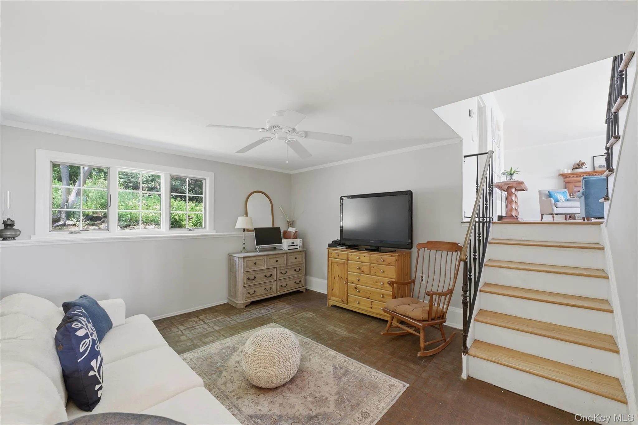 Lower-level family room featuring rich wood-finish flooring, a multi-pane window with exterior greenery, crown molding, and a ceiling fan Lower-level family room featuring rich wood-finish flooring, a multi-pane window with exterior greenery, crown molding, and a ceiling fan