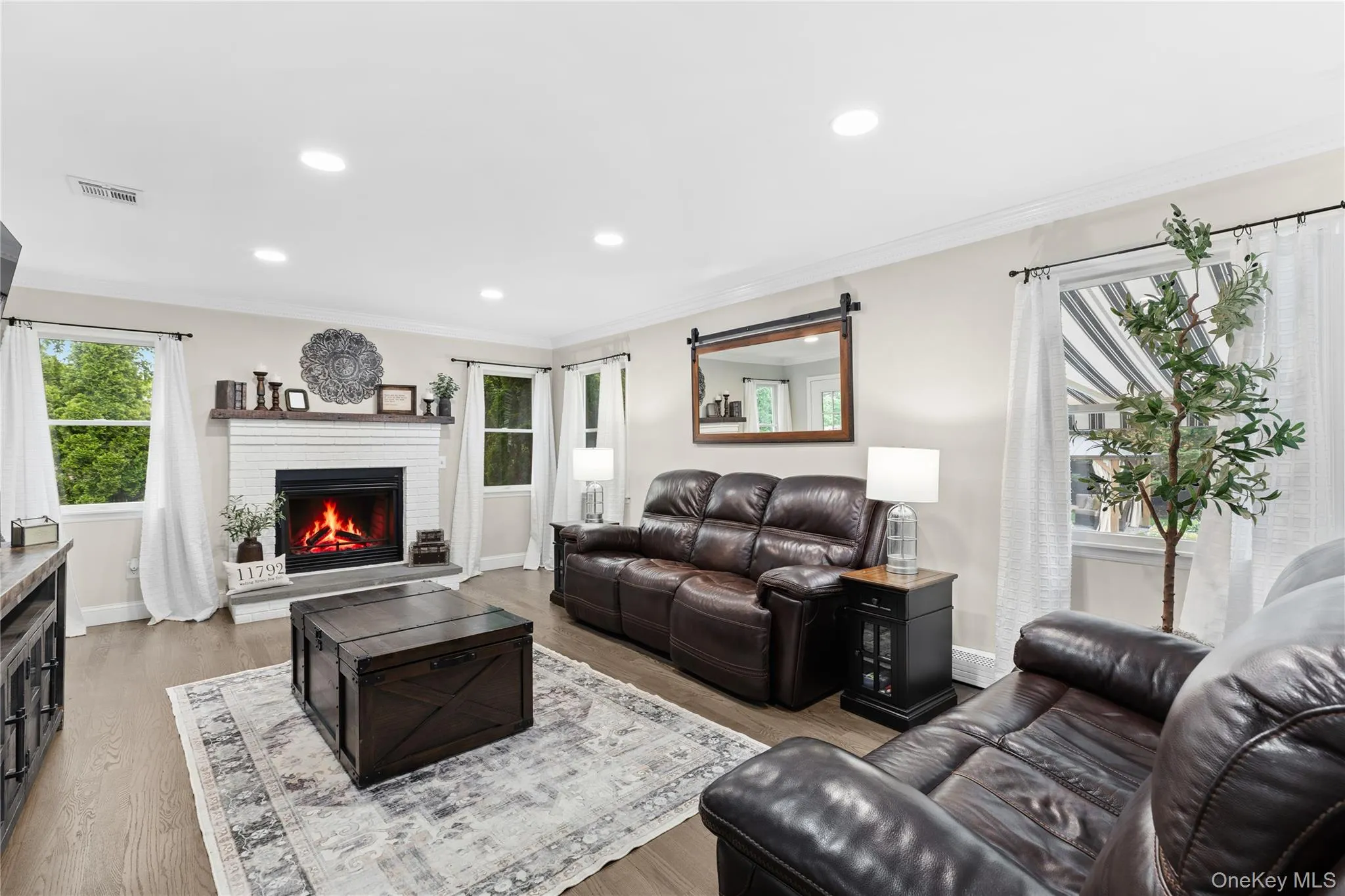 Spacious living area featuring a white brick fireplace with a dark wood mantel, recessed lighting, and wood-finish flooring Spacious living area featuring a white brick fireplace with a dark wood mantel, recessed lighting, and wood-finish flooring