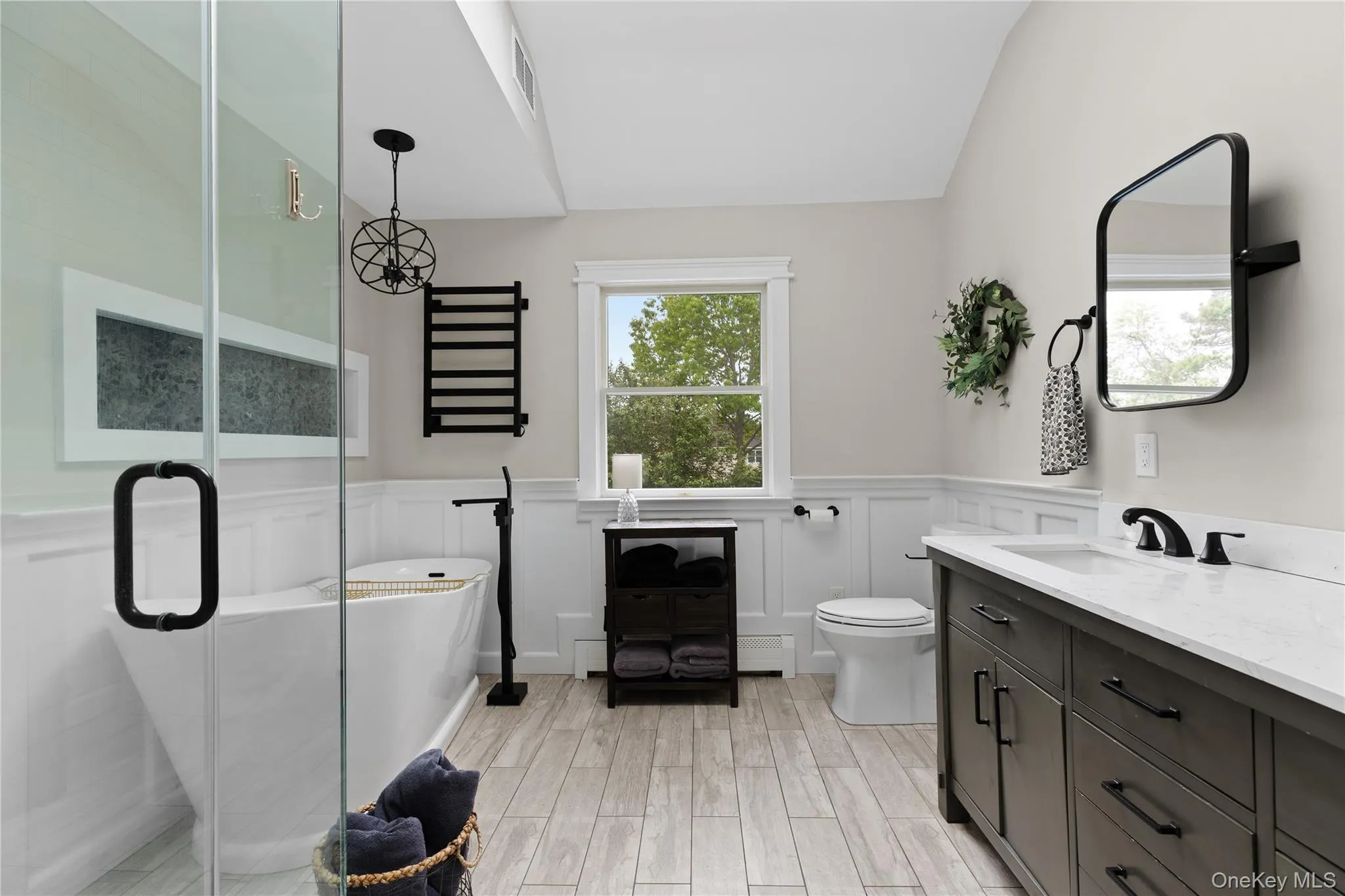Freestanding soaking tub with a floor-mounted fixture, glass-enclosed shower, wood-finish tile flooring, white wainscoting, and a double vanity with a light countertop Freestanding soaking tub with a floor-mounted fixture, glass-enclosed shower, wood-finish tile flooring, white wainscoting, and a double vanity with a light countertop