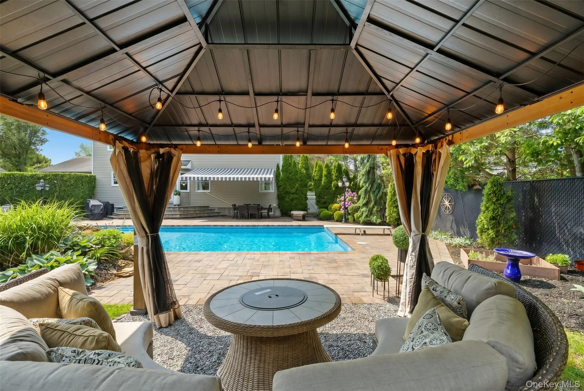 Gable-roof gazebo with string lighting, overlooking a paver patio and inground swimming pool Gable-roof gazebo with string lighting, overlooking a paver patio and inground swimming pool
