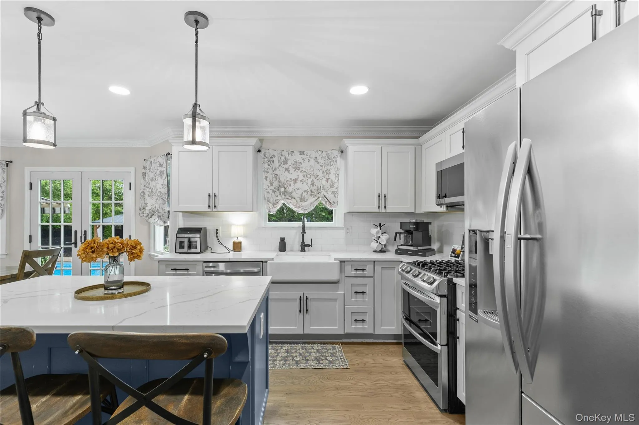 Kitchen featuring a central island with a white countertop, two pendant lights, white shaker-style cabinetry, stainless steel appliances, and wood-finish flooring Kitchen featuring a central island with a white countertop, two pendant lights, white shaker-style cabinetry, stainless steel appliances, and wood-finish flooring