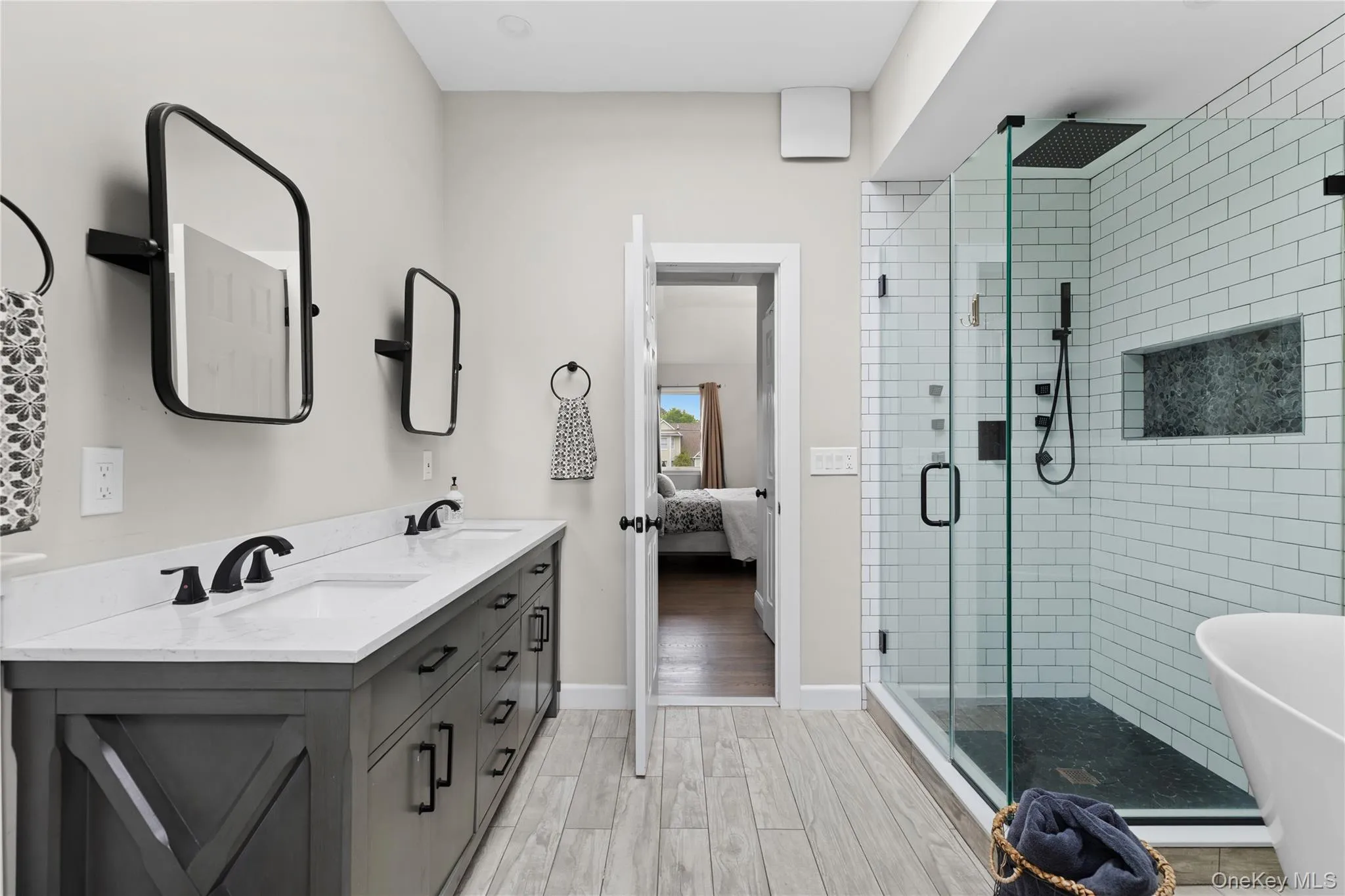 Bathroom featuring a dual vanity with white countertops, a glass-enclosed shower with white subway tile, black fixtures, and wood-finish flooring Bathroom featuring a dual vanity with white countertops, a glass-enclosed shower with white subway tile, black fixtures, and wood-finish flooring