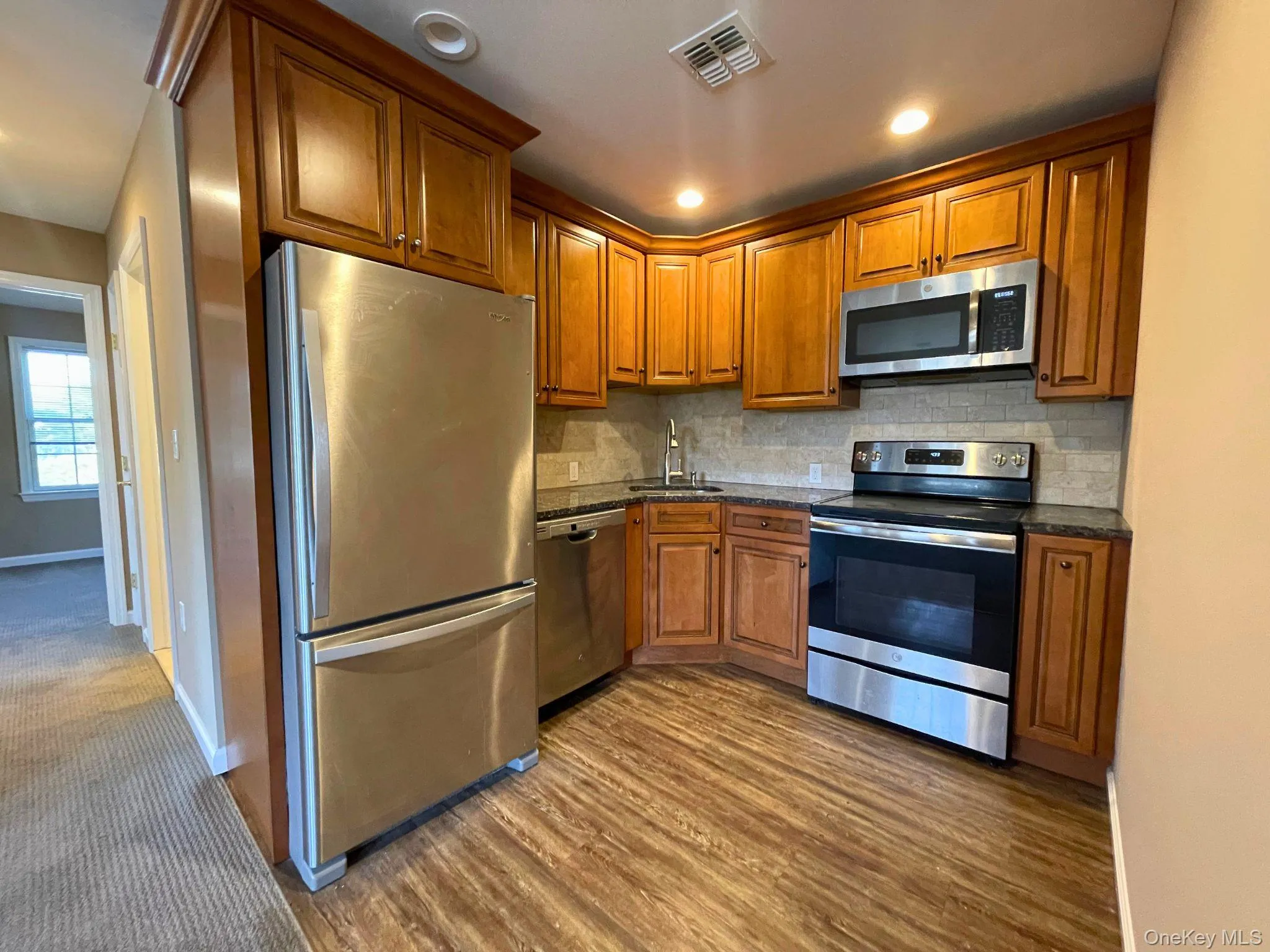Kitchen featuring wood-finish flooring, full-height cabinetry with crown molding, stainless steel appliances, granite-style countertops, and a stone-look tile backsplash Kitchen featuring wood-finish flooring, full-height cabinetry with crown molding, stainless steel appliances, granite-style countertops, and a stone-look tile backsplash