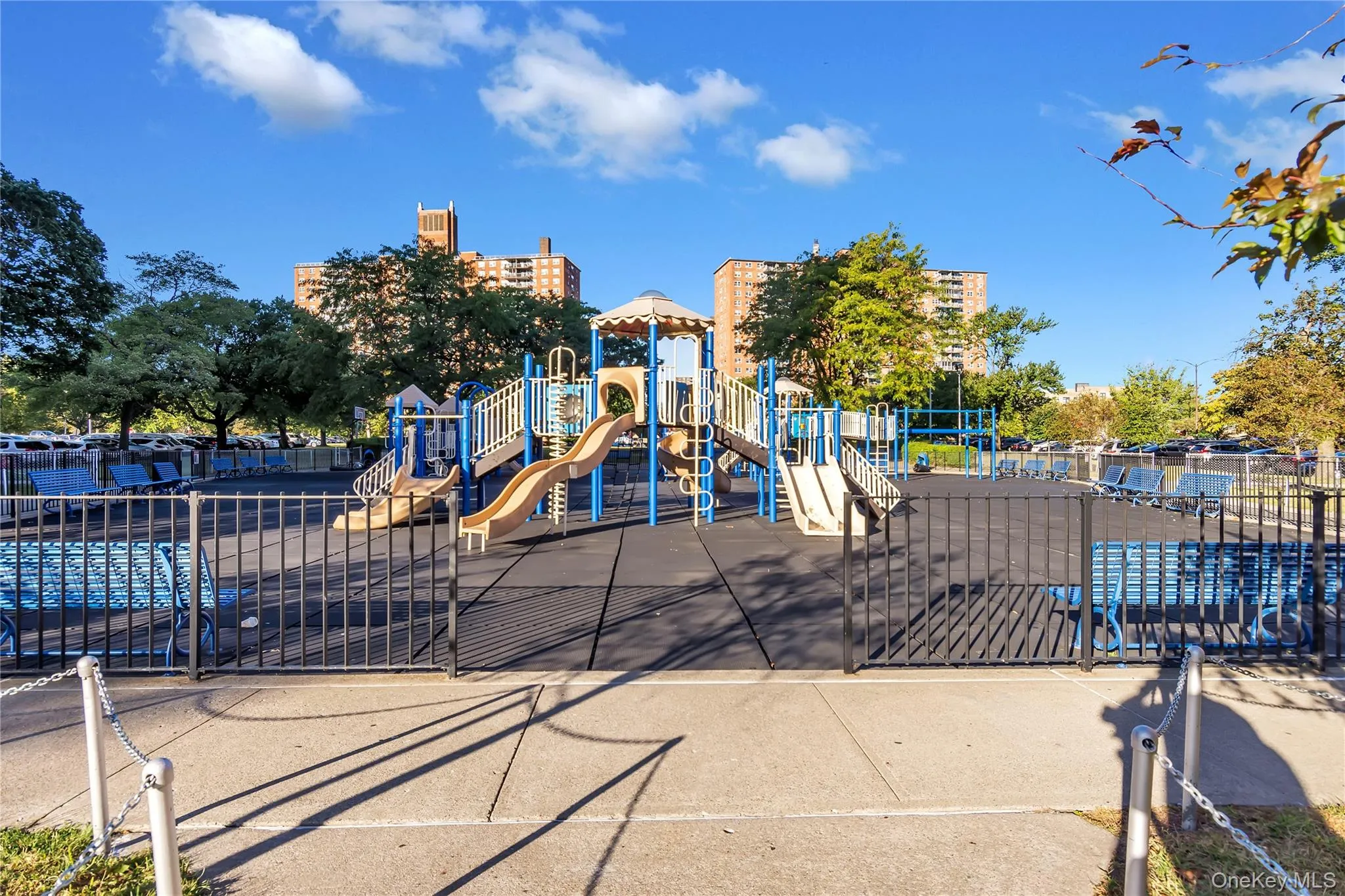Gated outdoor play area featuring multiple slides and climbing structures, with a rubberized surface and surrounding park benches Gated outdoor play area featuring multiple slides and climbing structures, with a rubberized surface and surrounding park benches