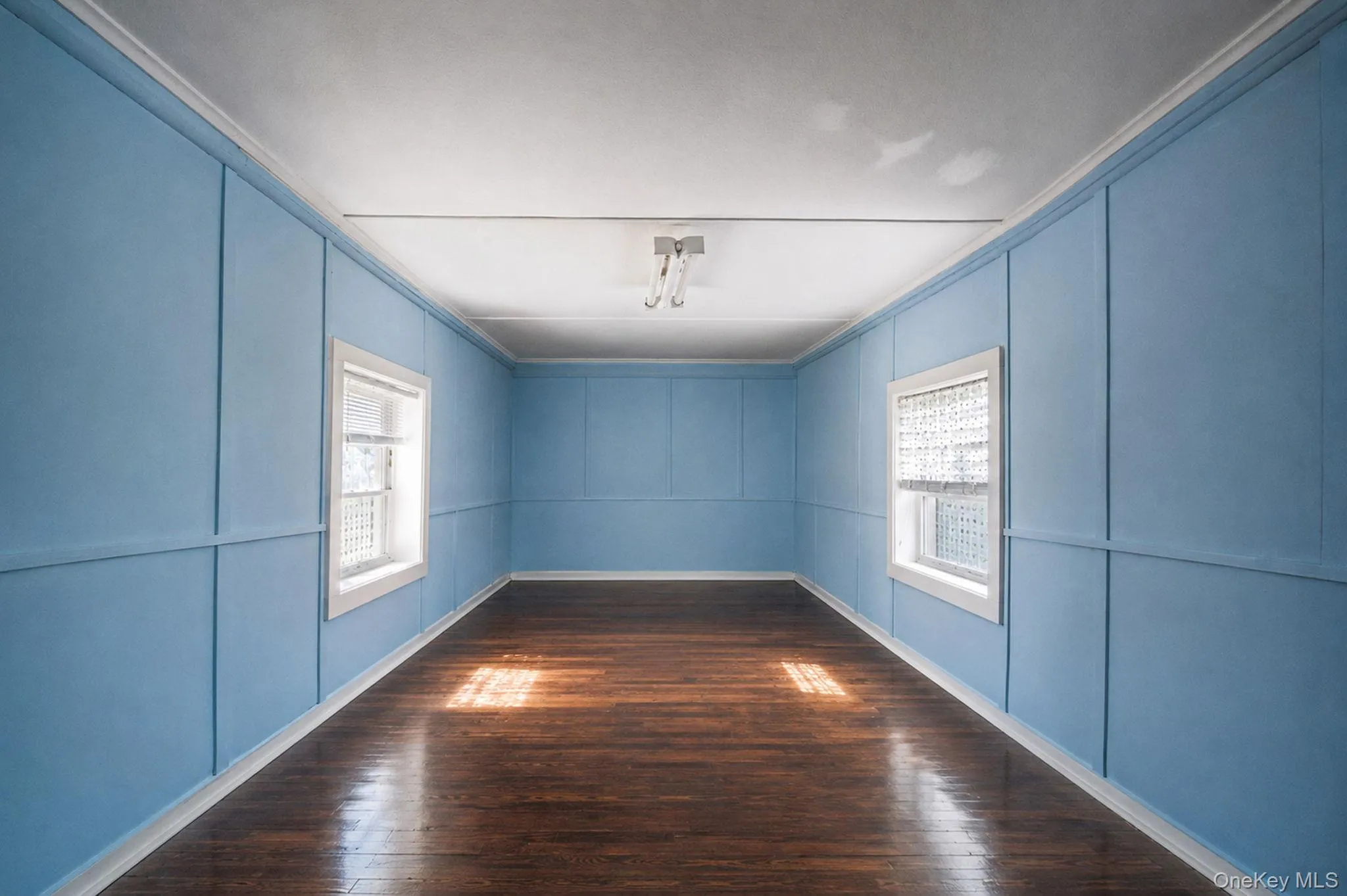 Room featuring wood-finish flooring, two windows with white trim, and light blue wall paneling Room featuring wood-finish flooring, two windows with white trim, and light blue wall paneling