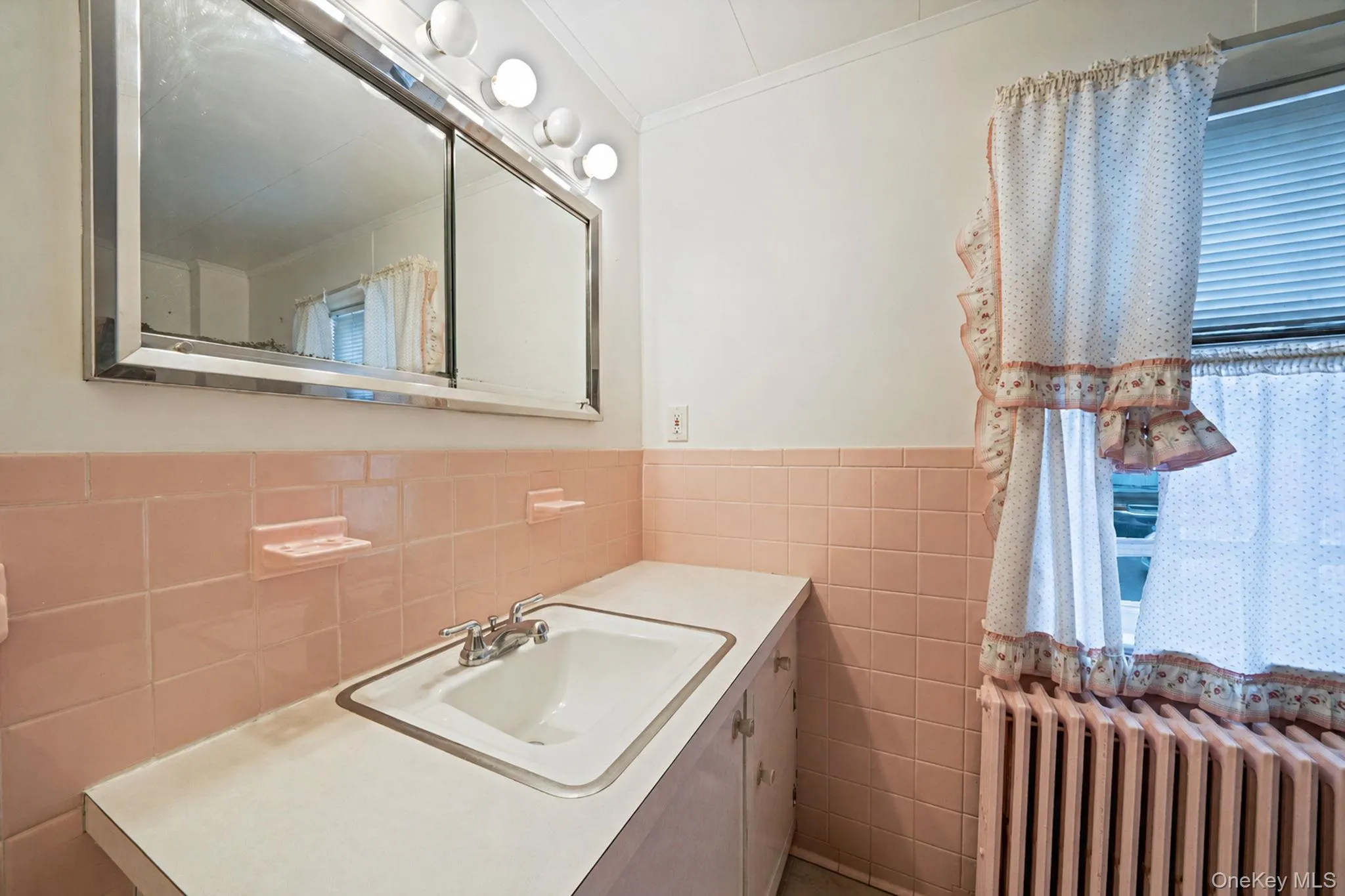 Bathroom featuring pink tiled wainscoting, a wall-mounted vanity mirror with integrated lighting, a drop-in sink with a chrome faucet, r Bathroom featuring pink tiled wainscoting, a wall-mounted vanity mirror with integrated lighting, a drop-in sink with a chrome faucet, r