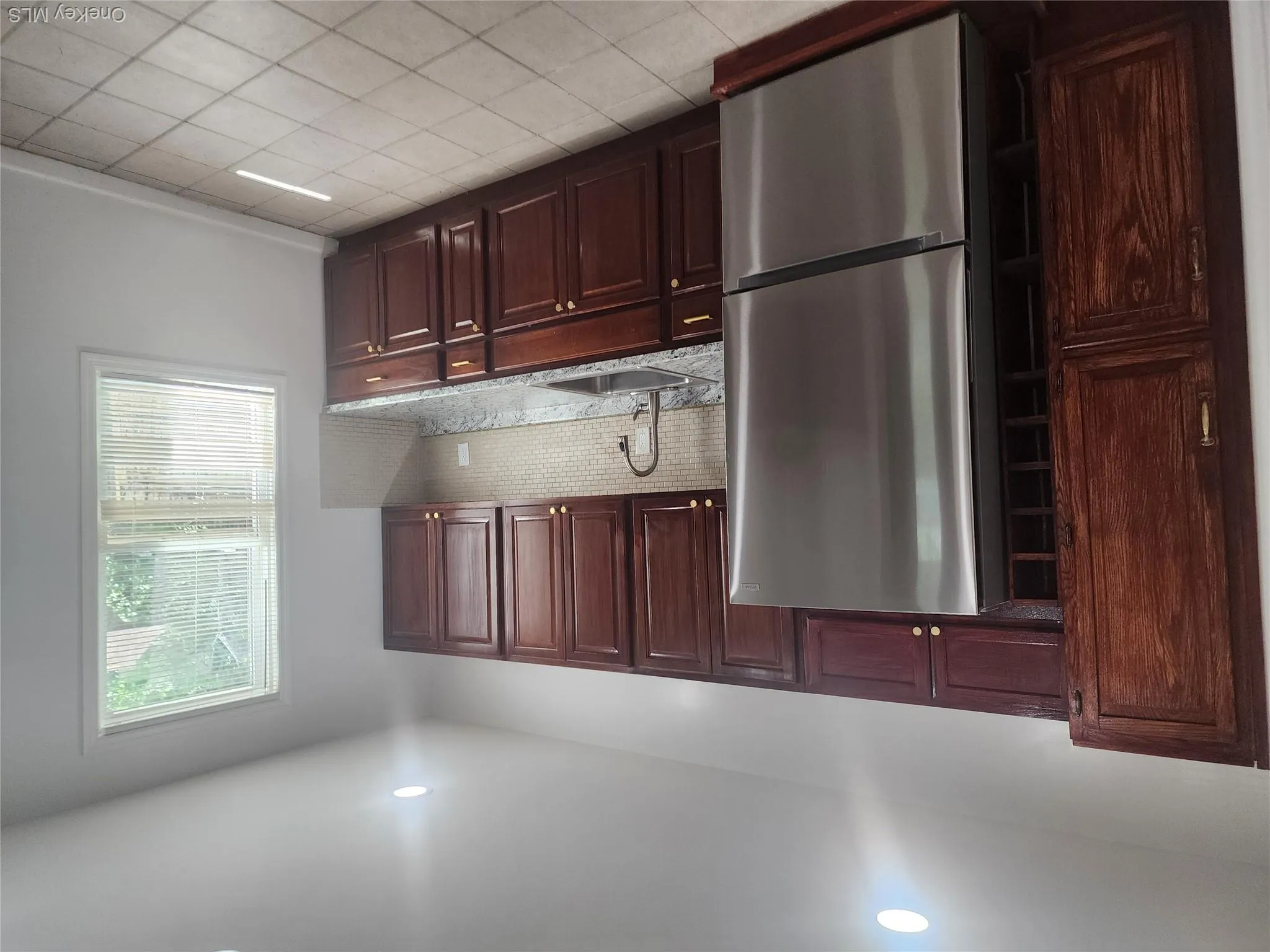 Kitchen featuring dark wood-finish cabinetry with gold-tone hardware, a stainless steel refrigerator, granite-style countertops, and a mosaic tile backsplash with tile floor. Kitchen featuring dark wood-finish cabinetry with gold-tone hardware, a stainless steel refrigerator, granite-style countertops, and a mosaic tile backsplash with tile floor.
