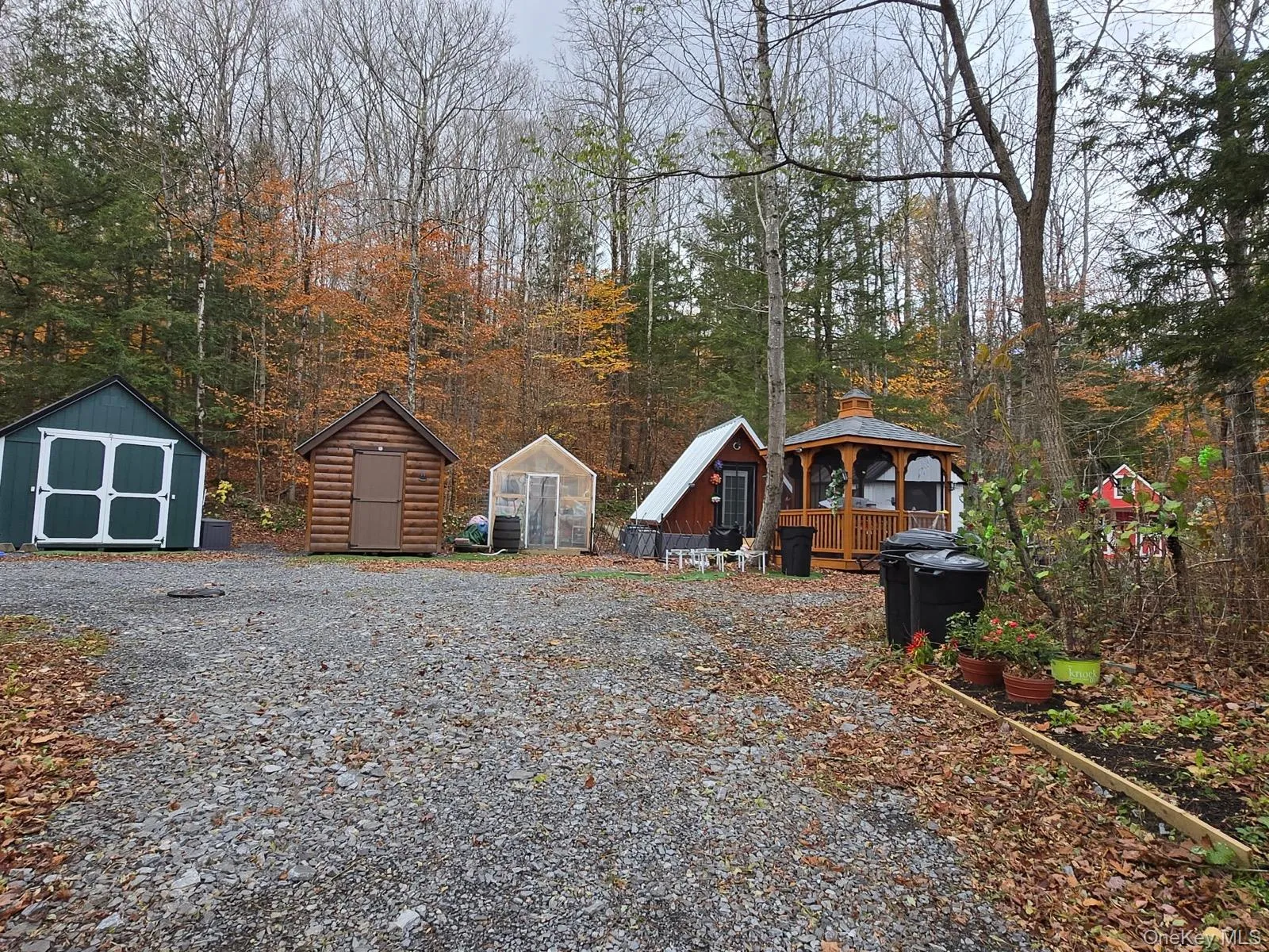 Gravel driveway area featuring multiple outbuildings, including a green storage shed, a log cabin-style shed, and a greenhouse Gravel driveway area featuring multiple outbuildings, including a green storage shed, a log cabin-style shed, and a greenhouse