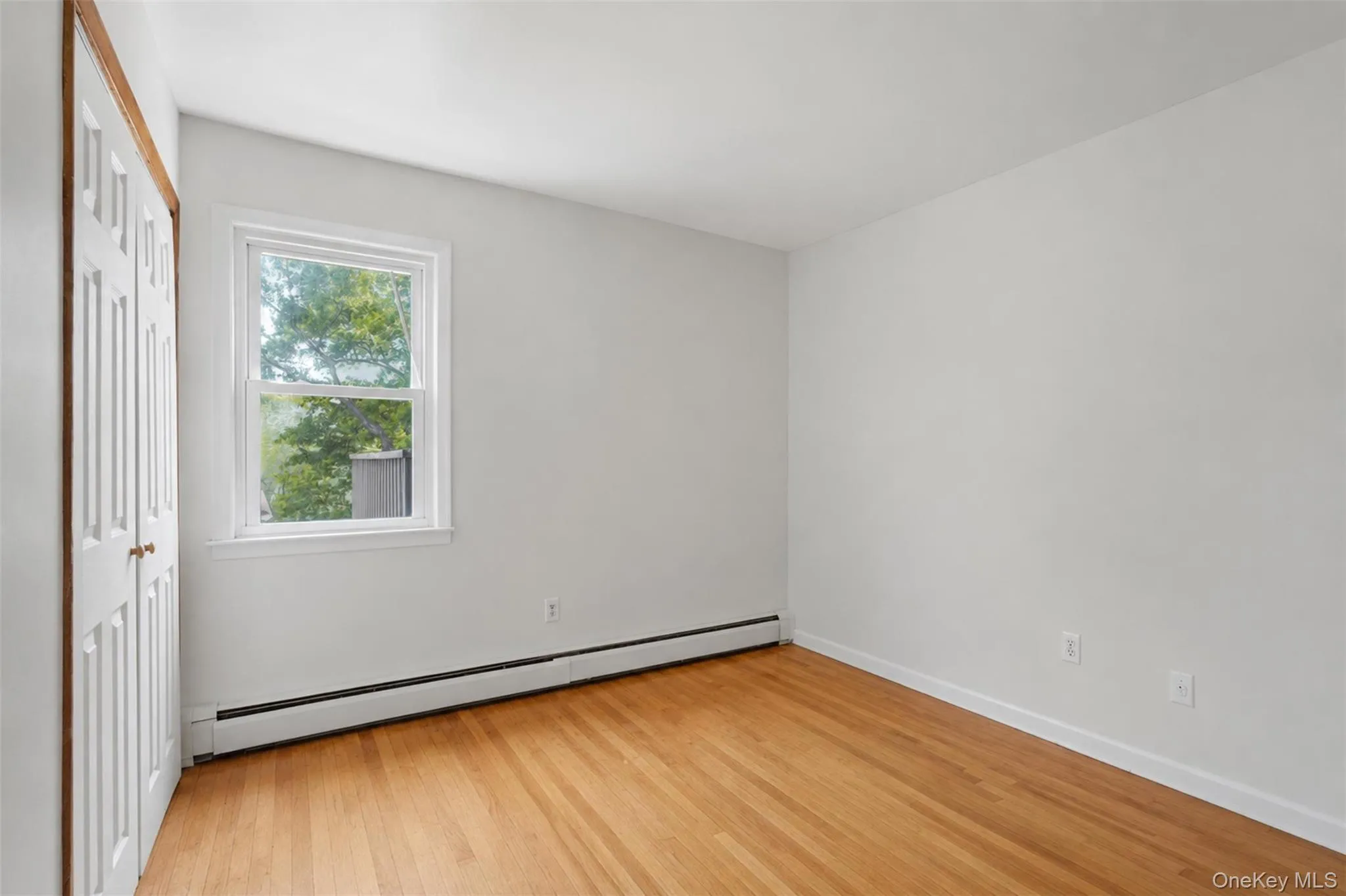 Room featuring wood-finish flooring, a double-hung window, white walls, and bi-fold closet doors Room featuring wood-finish flooring, a double-hung window, white walls, and bi-fold closet doors