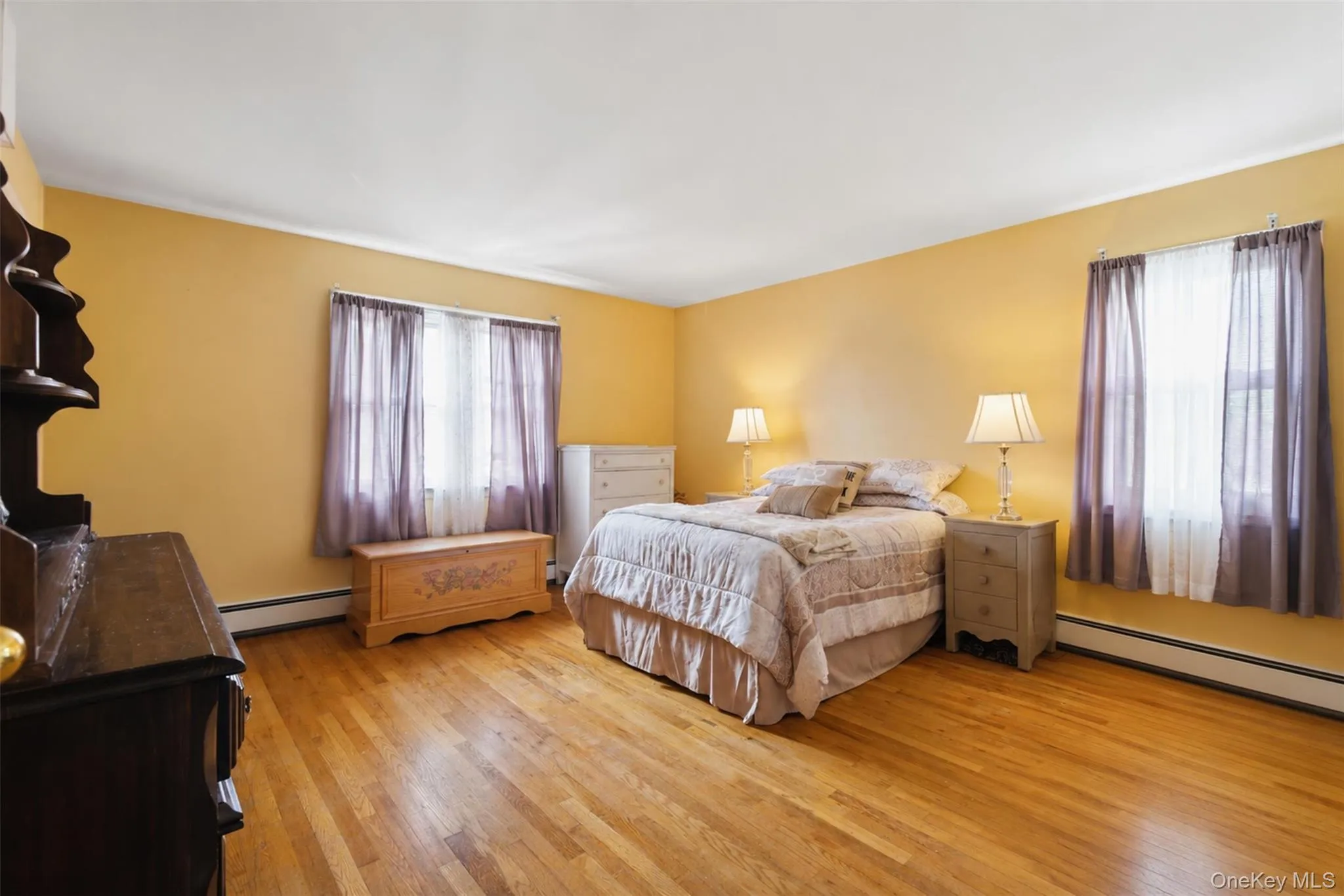 Bright room featuring hardwood flooring, light yellow walls, white baseboards, a window with sheer and opaque curtains, and a white ceiling Bright room featuring hardwood flooring, light yellow walls, white baseboards, a window with sheer and opaque curtains, and a white ceiling