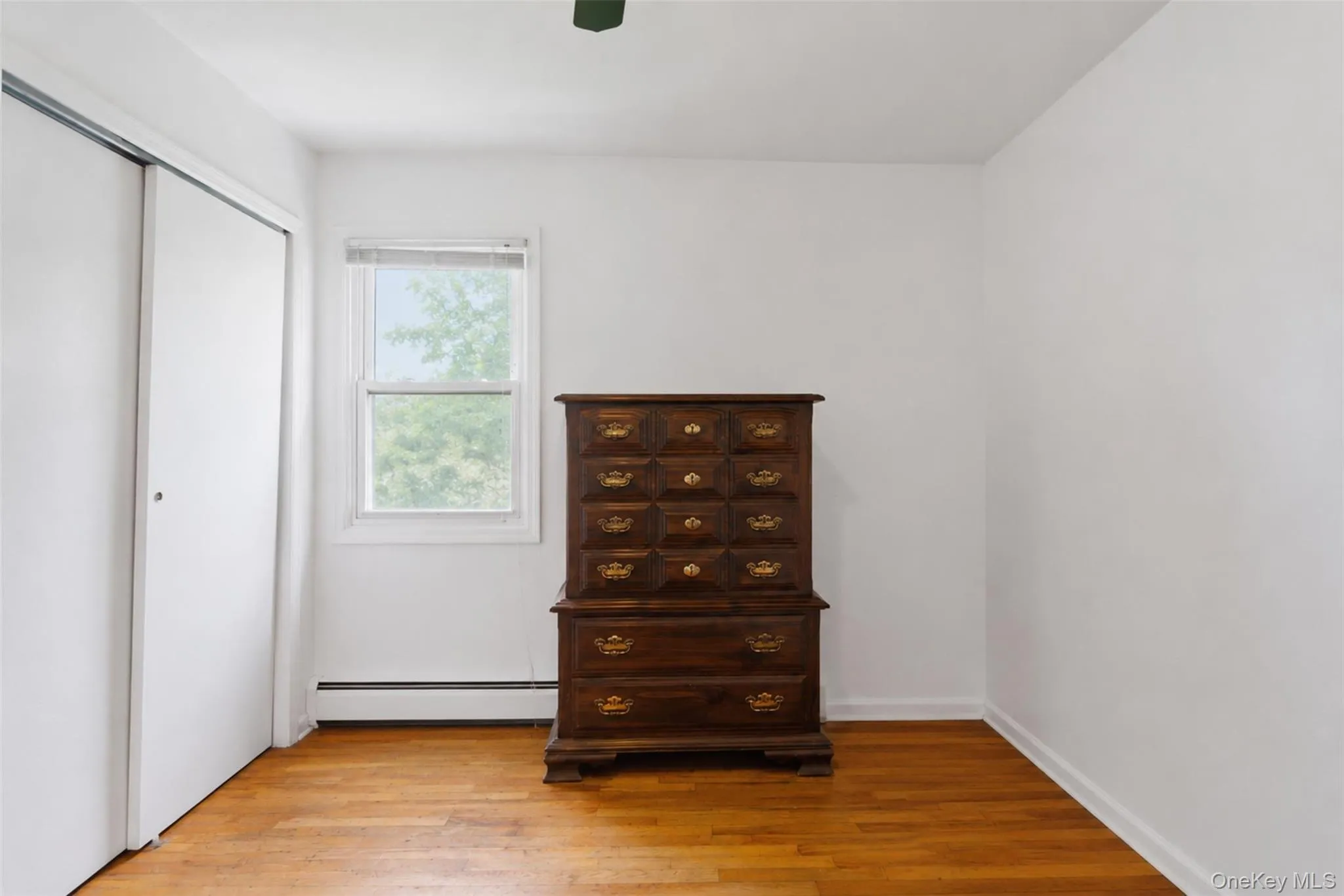 Light-filled room featuring wood-finish flooring, white walls, and a window with exterior tree views Light-filled room featuring wood-finish flooring, white walls, and a window with exterior tree views