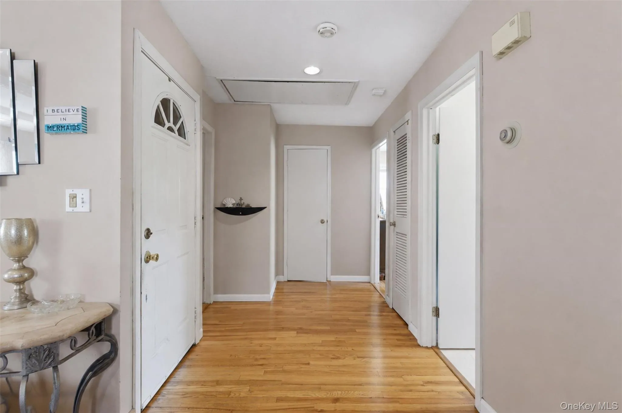 Hardwood flooring extends throughout this interior hallway, featuring multiple white paneled doors and an attic access panel Hardwood flooring extends throughout this interior hallway, featuring multiple white paneled doors and an attic access panel