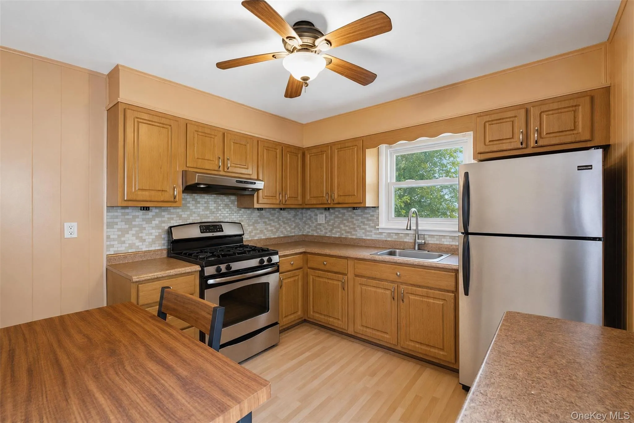 Kitchen featuring wood-finish cabinetry, stainless steel appliances, mosaic tile backsplash, and a ceiling fan with integrated lighting Kitchen featuring wood-finish cabinetry, stainless steel appliances, mosaic tile backsplash, and a ceiling fan with integrated lighting