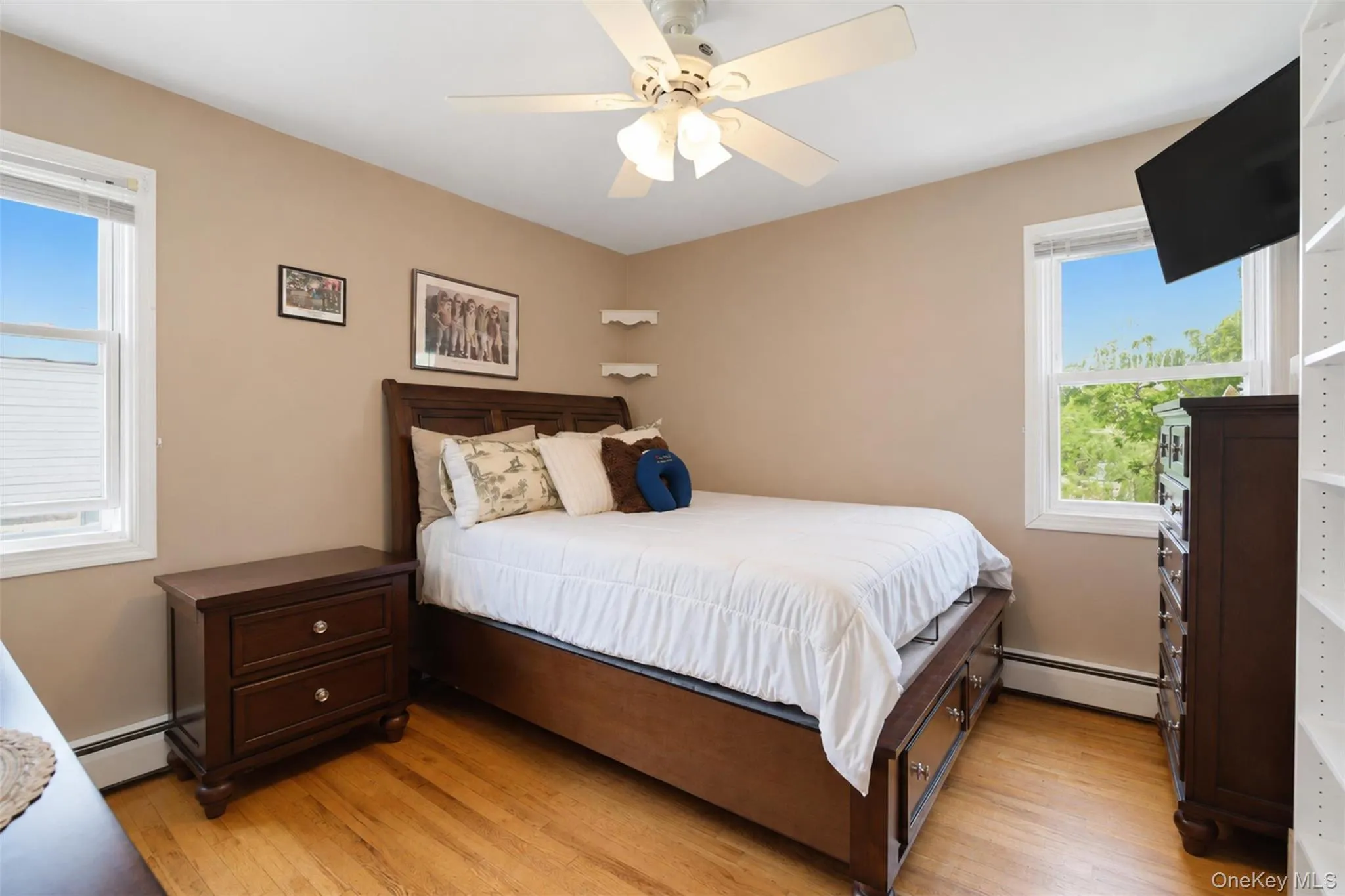 Hardwood flooring extends throughout this room, featuring two windows, a ceiling fan with integrated lighting, and neutral wall tones Hardwood flooring extends throughout this room, featuring two windows, a ceiling fan with integrated lighting, and neutral wall tones
