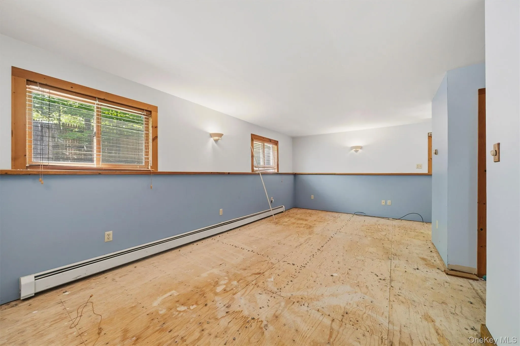 Spacious room featuring plywood subflooring, multiple windows with wood trim, and wall-mounted sconce lighting Spacious room featuring plywood subflooring, multiple windows with wood trim, and wall-mounted sconce lighting