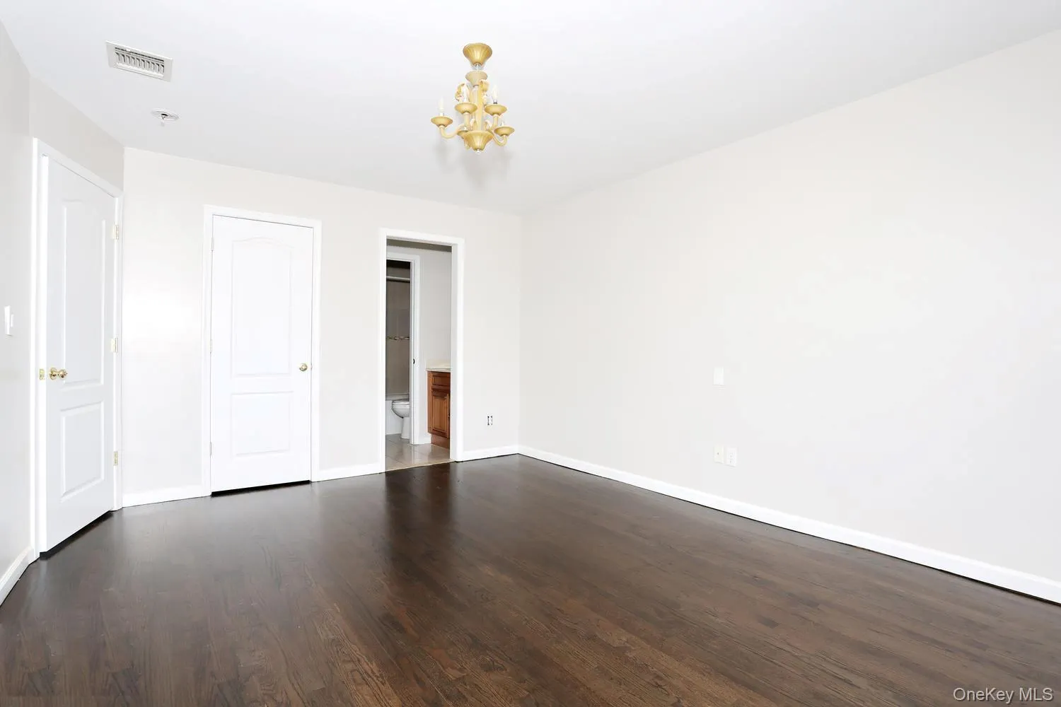 Spacious room featuring dark wood-finish flooring, white baseboards, and a gold-toned ceiling light fixture Spacious room featuring dark wood-finish flooring, white baseboards, and a gold-toned ceiling light fixture