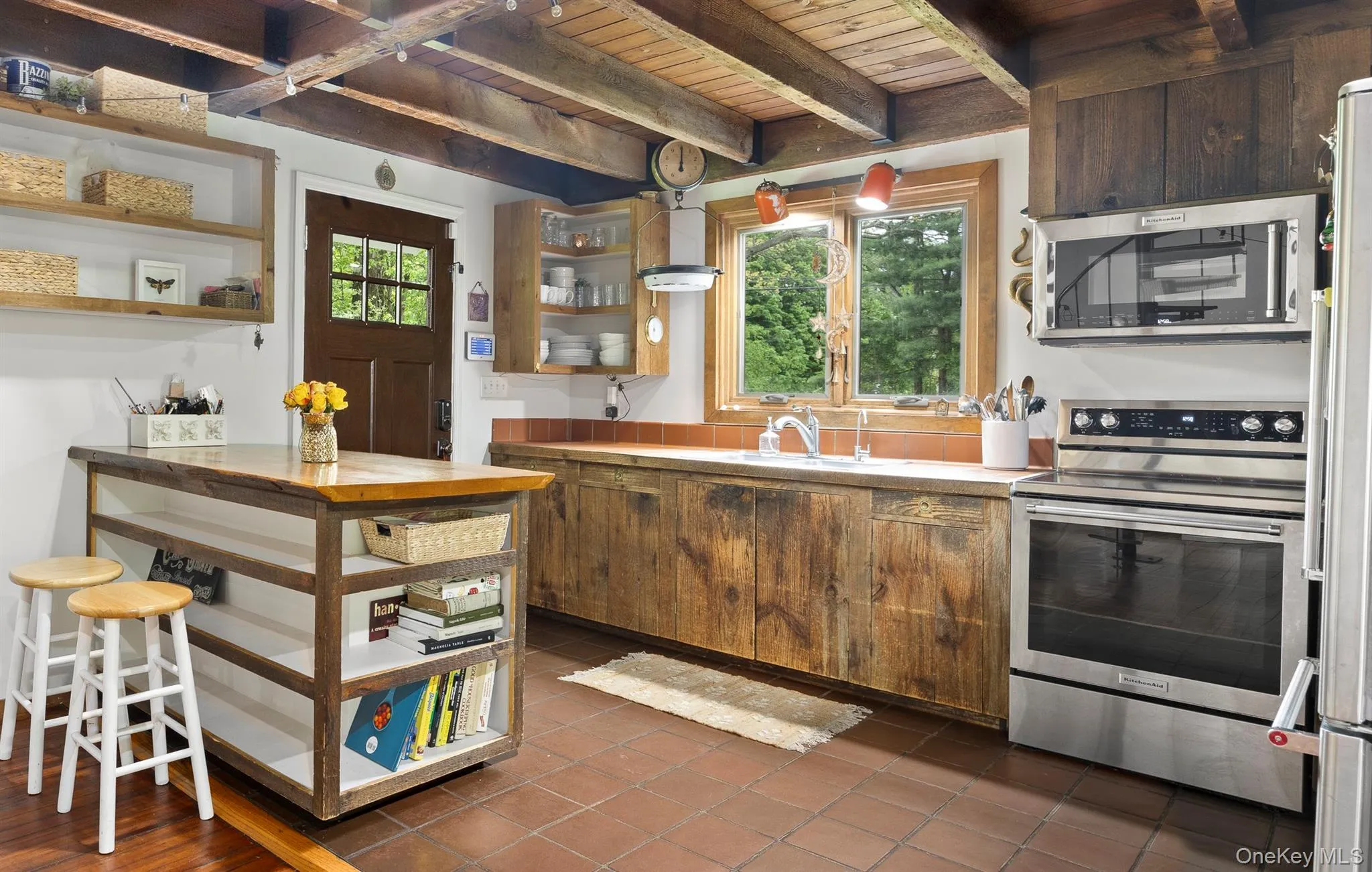 Kitchen featuring exposed wood beam ceiling, rustic wood cabinetry, stainless steel appliances, a center island with open shelving, and terracotta tile flooring Kitchen featuring exposed wood beam ceiling, rustic wood cabinetry, stainless steel appliances, a center island with open shelving, and terracotta tile flooring