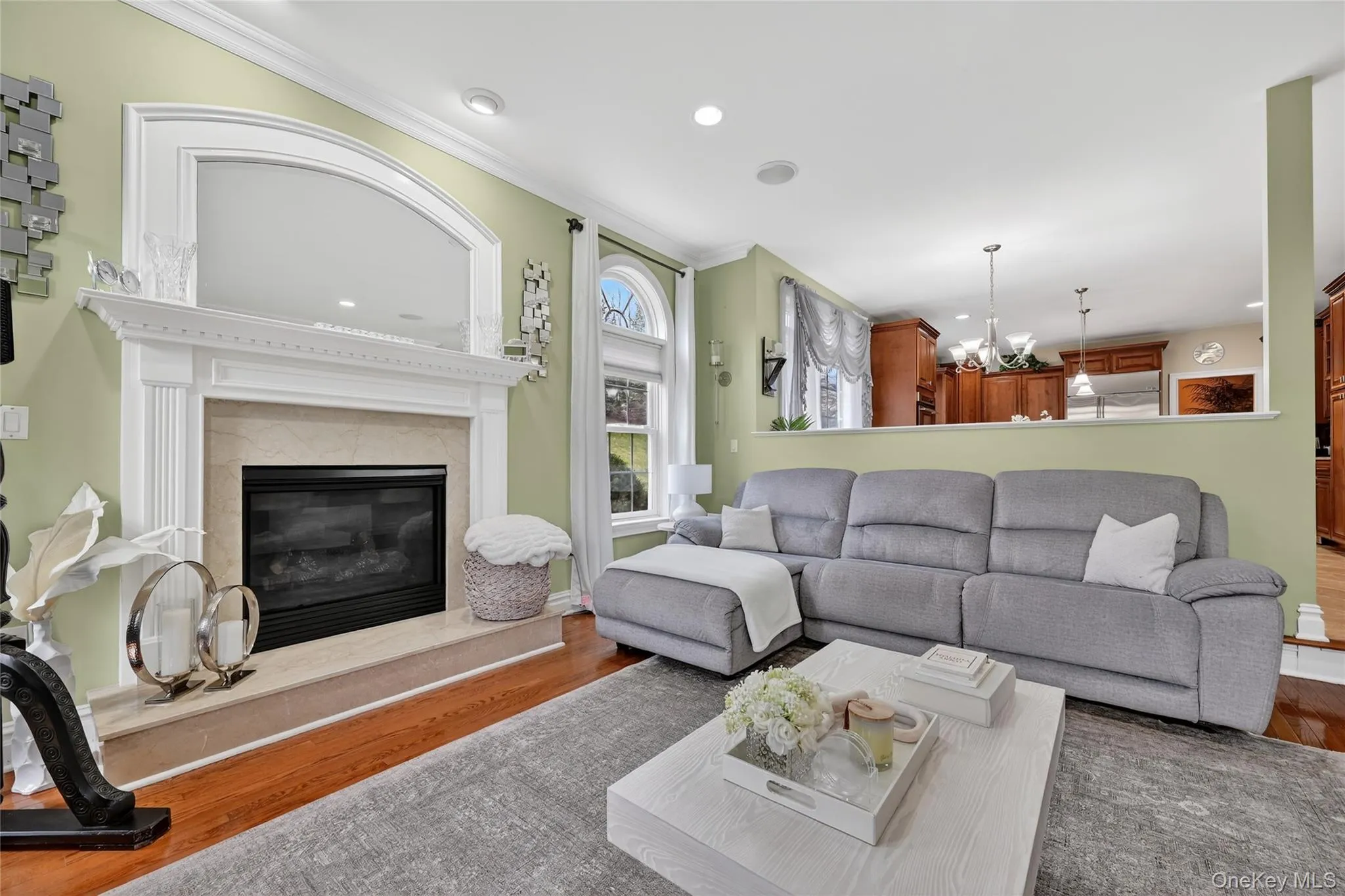 Living area featuring a white mantel fireplace with a neutral stone surround, wood-finish flooring, recessed lighting, and an arched window Living area featuring a white mantel fireplace with a neutral stone surround, wood-finish flooring, recessed lighting, and an arched window