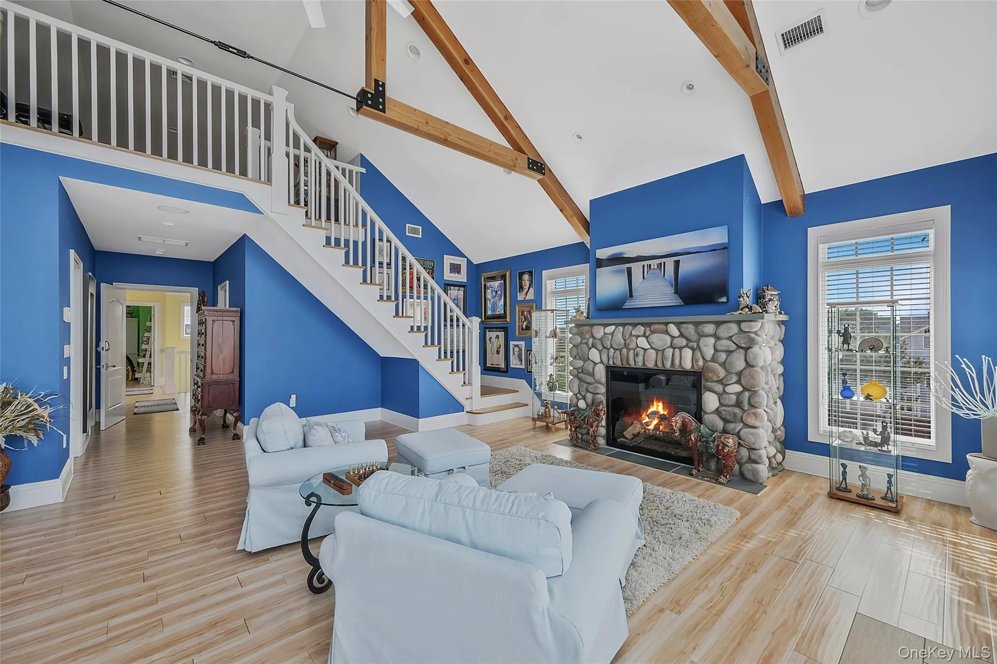 Spacious primary sitting area featuring a stone-clad gas fireplace, vaulted ceiling with exposed wood beams, a prominent staircase with white balusters, wood-finish flooring, and large windows with blinds Spacious primary sitting area featuring a stone-clad gas fireplace, vaulted ceiling with exposed wood beams, a prominent staircase with white balusters, wood-finish flooring, and large windows with blinds
