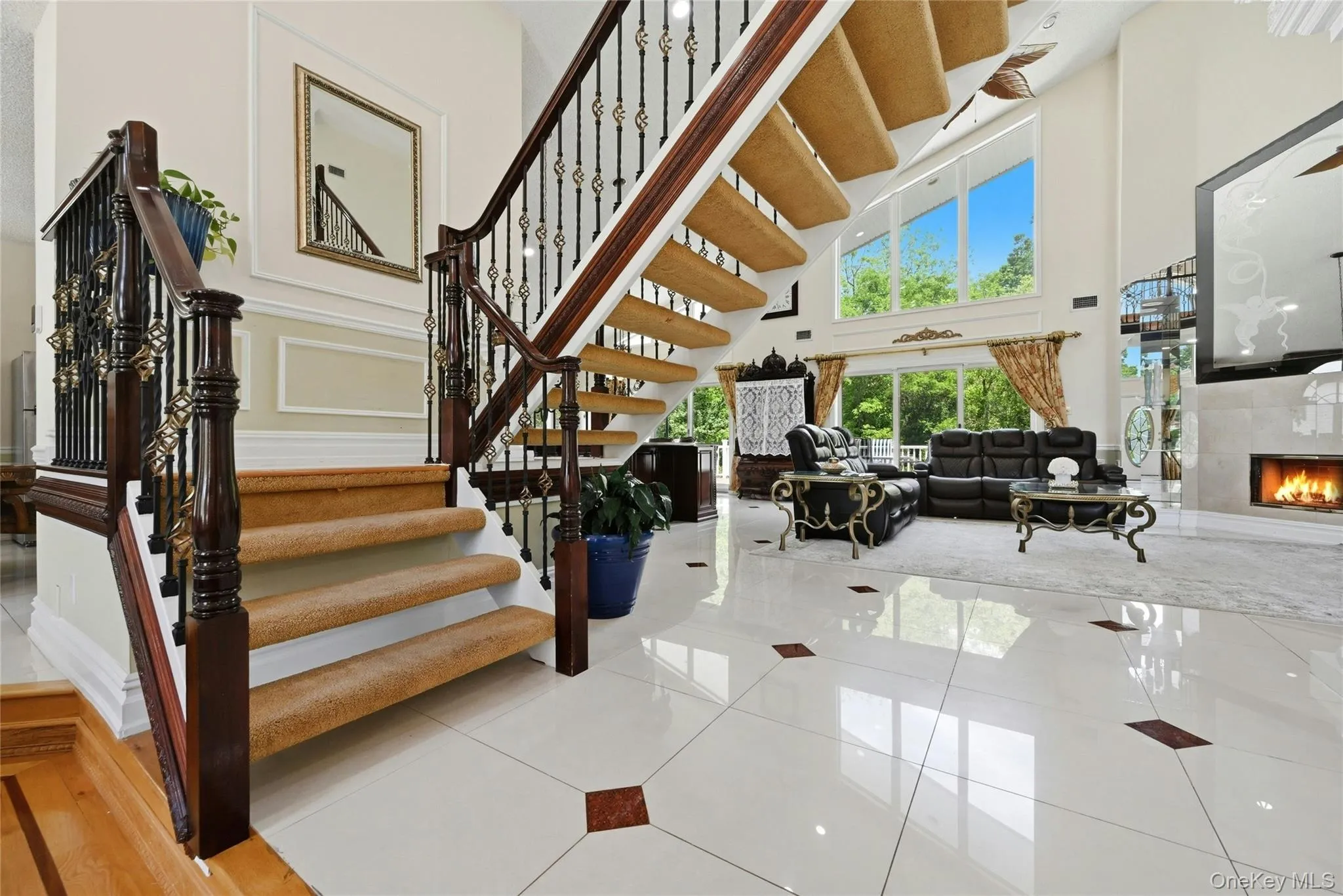 Grand two-story foyer featuring a carved wood banister with ornate metal spindles, white tile flooring with accent inlays, and a wall of windows offering exterior views Grand two-story foyer featuring a carved wood banister with ornate metal spindles, white tile flooring with accent inlays, and a wall of windows offering exterior views