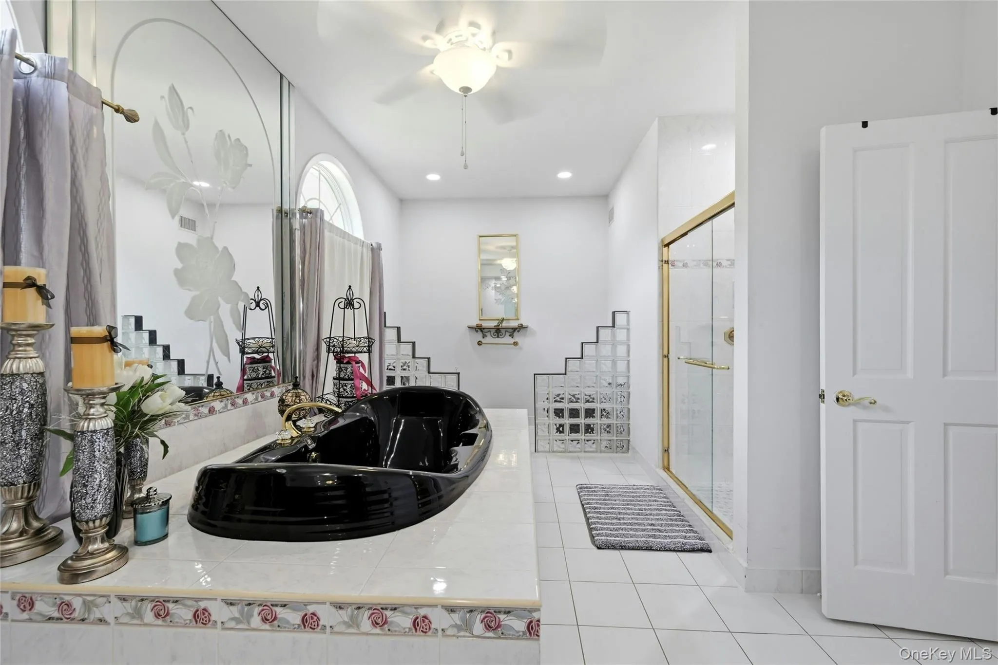 Spacious bathroom featuring a black soaking tub, a large mirrored wall with etched detailing, a glass-enclosed shower with gold-tone framing, a tiered glass block partition, and white tile flooring Spacious bathroom featuring a black soaking tub, a large mirrored wall with etched detailing, a glass-enclosed shower with gold-tone framing, a tiered glass block partition, and white tile flooring