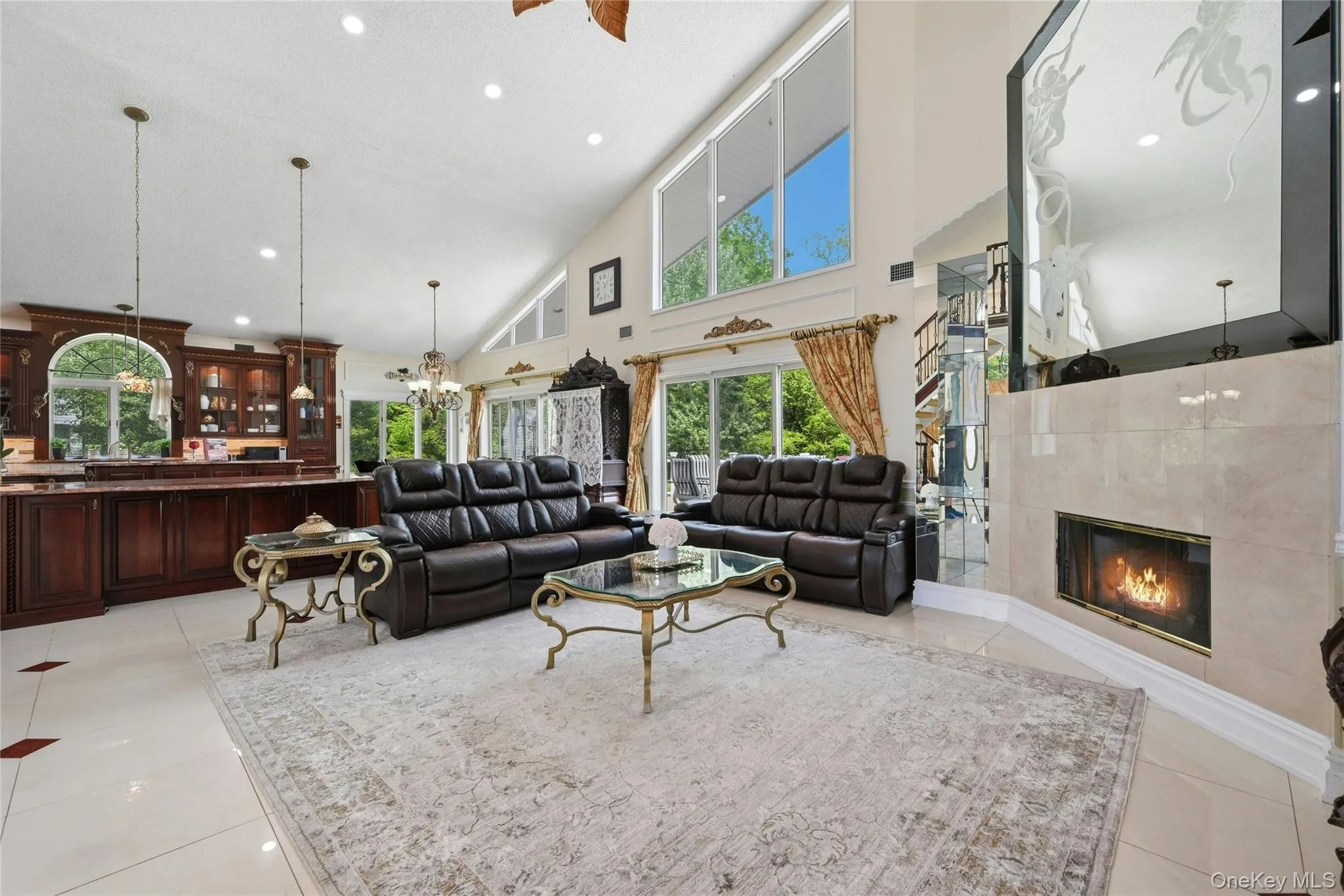 Spacious living area featuring a vaulted ceiling with recessed lighting, a built-in fireplace with a stone surround, large windows, and an adjacent kitchen with dark wood cabinetry and a peninsula Spacious living area featuring a vaulted ceiling with recessed lighting, a built-in fireplace with a stone surround, large windows, and an adjacent kitchen with dark wood cabinetry and a peninsula