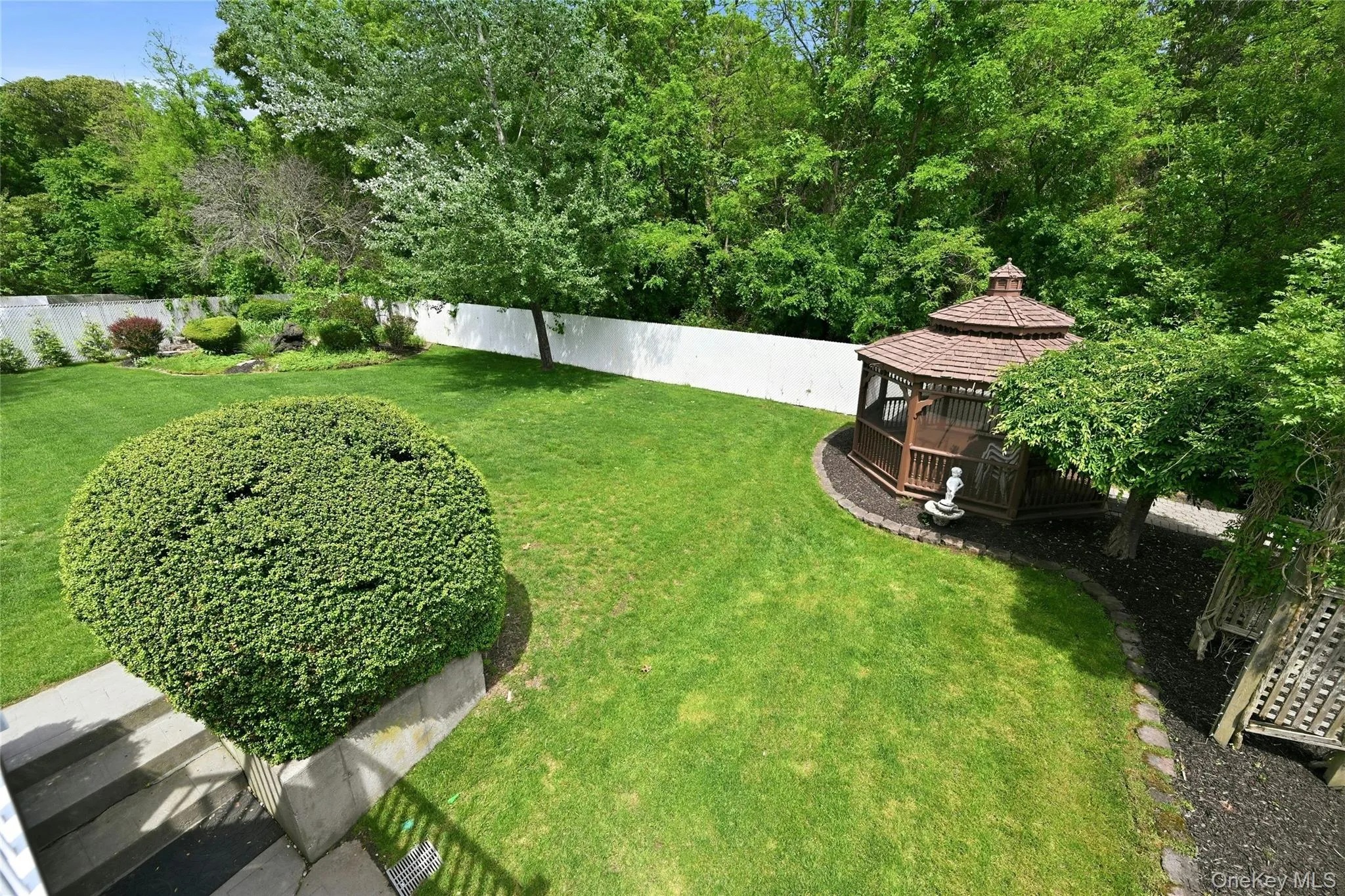 Expansive green lawn featuring a wooden gazebo with a shingle roof, surrounded by mature trees and a white privacy fence Expansive green lawn featuring a wooden gazebo with a shingle roof, surrounded by mature trees and a white privacy fence