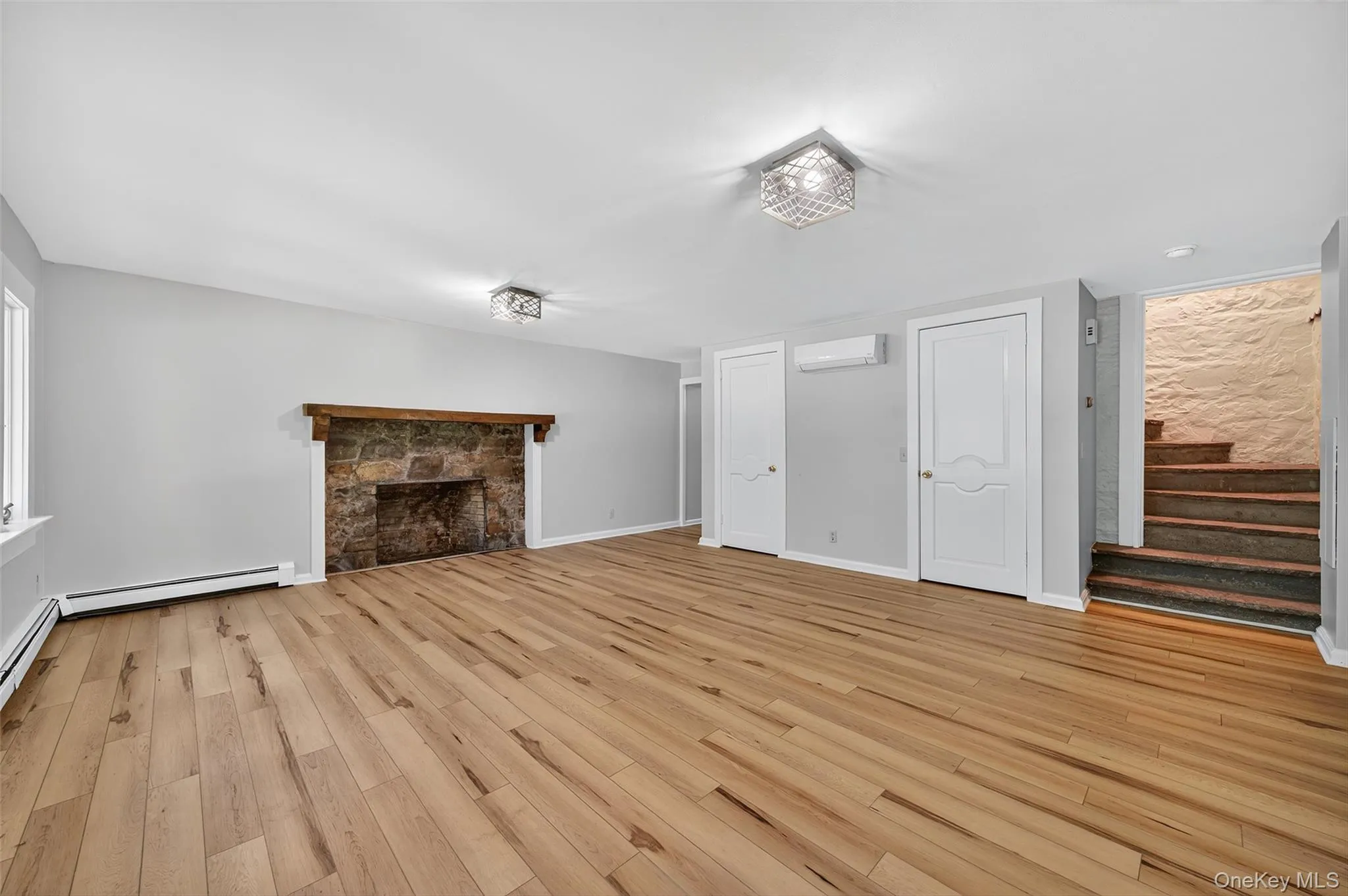 Unfurnished with stone stairway to second floor, wood-style flooring, decorative stone fireplace, baseboard heating, and a wall mounted HVAC mini-split Unfurnished with stone stairway to second floor, wood-style flooring, decorative stone fireplace, baseboard heating, and a wall mounted HVAC mini-split