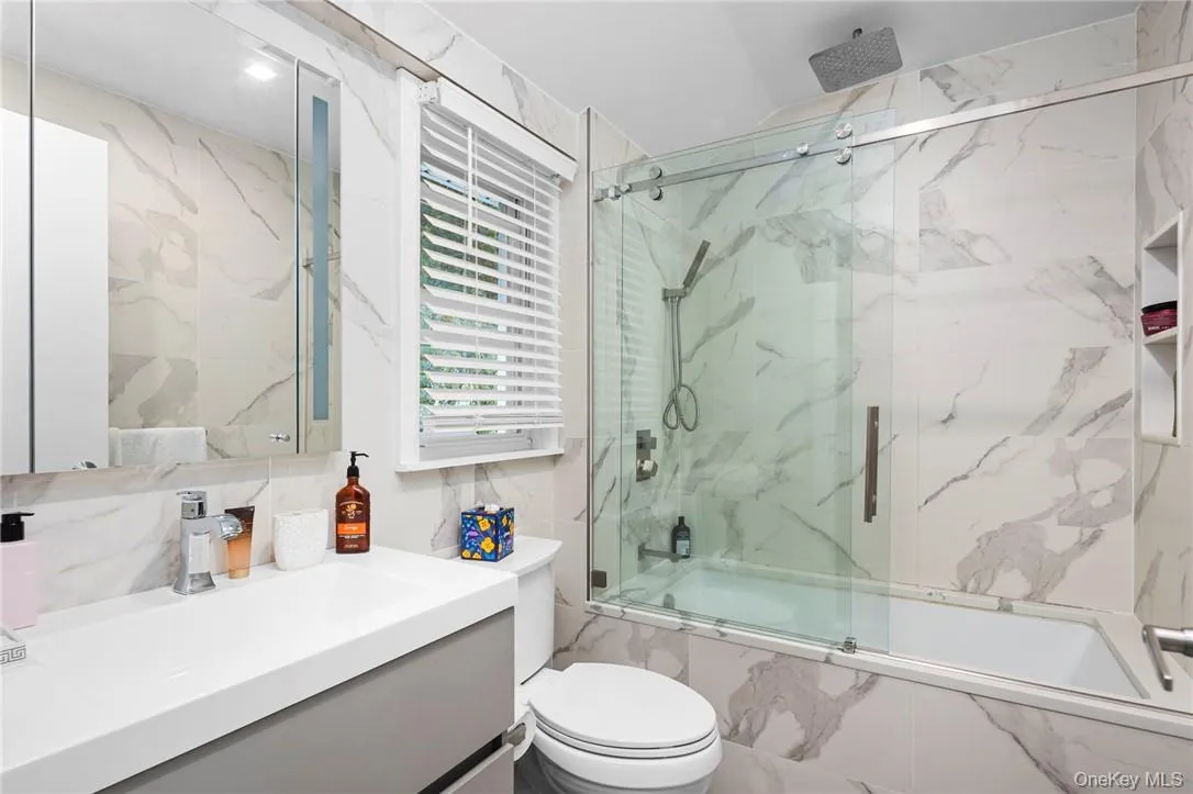 Full bathroom with tile walls, bath / shower combo with glass door, toilet, and vanity Full bathroom with tile walls, bath / shower combo with glass door, toilet, and vanity