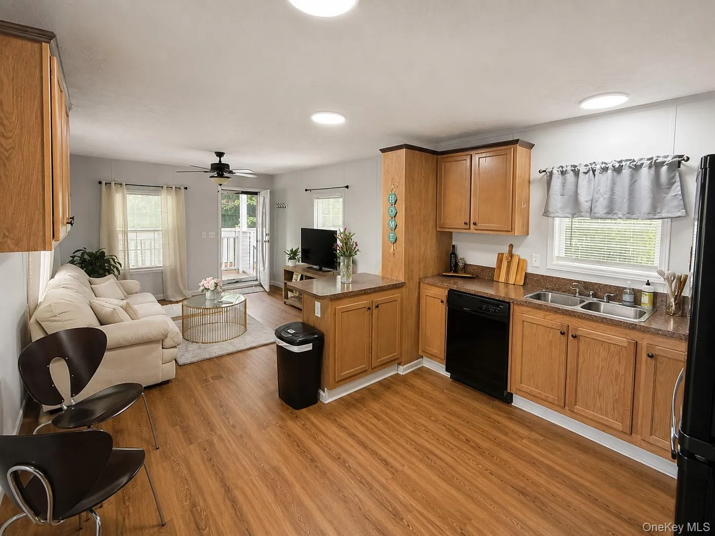 Open-concept living area featuring wood-finish flooring, a kitchen with wood cabinetry and countertop space, a stainless steel double sink, and a black dishwasher Open-concept living area featuring wood-finish flooring, a kitchen with wood cabinetry and countertop space, a stainless steel double sink, and a black dishwasher