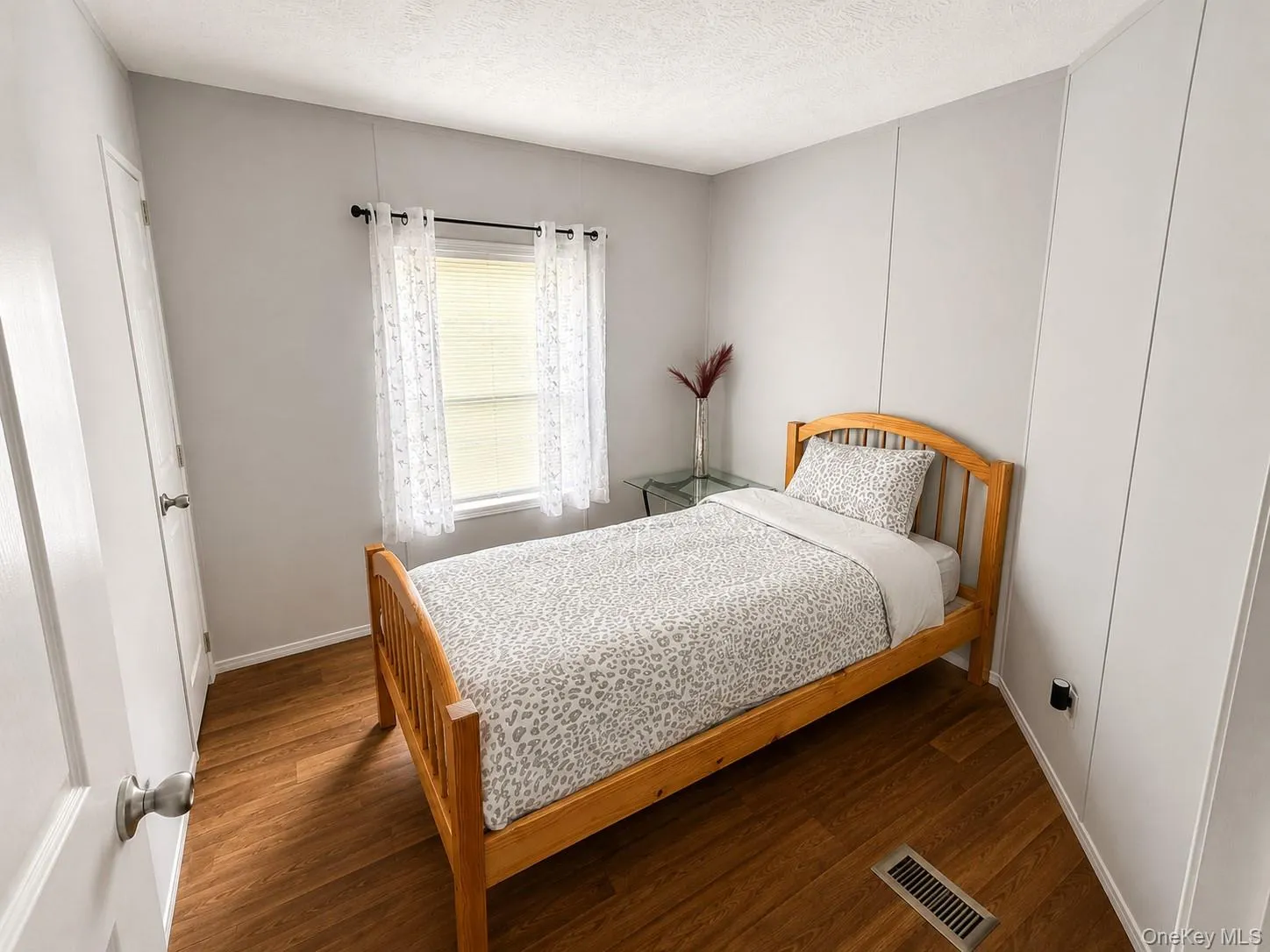 Room featuring wood-finish flooring, light gray wall panels, and a window with white blinds Room featuring wood-finish flooring, light gray wall panels, and a window with white blinds