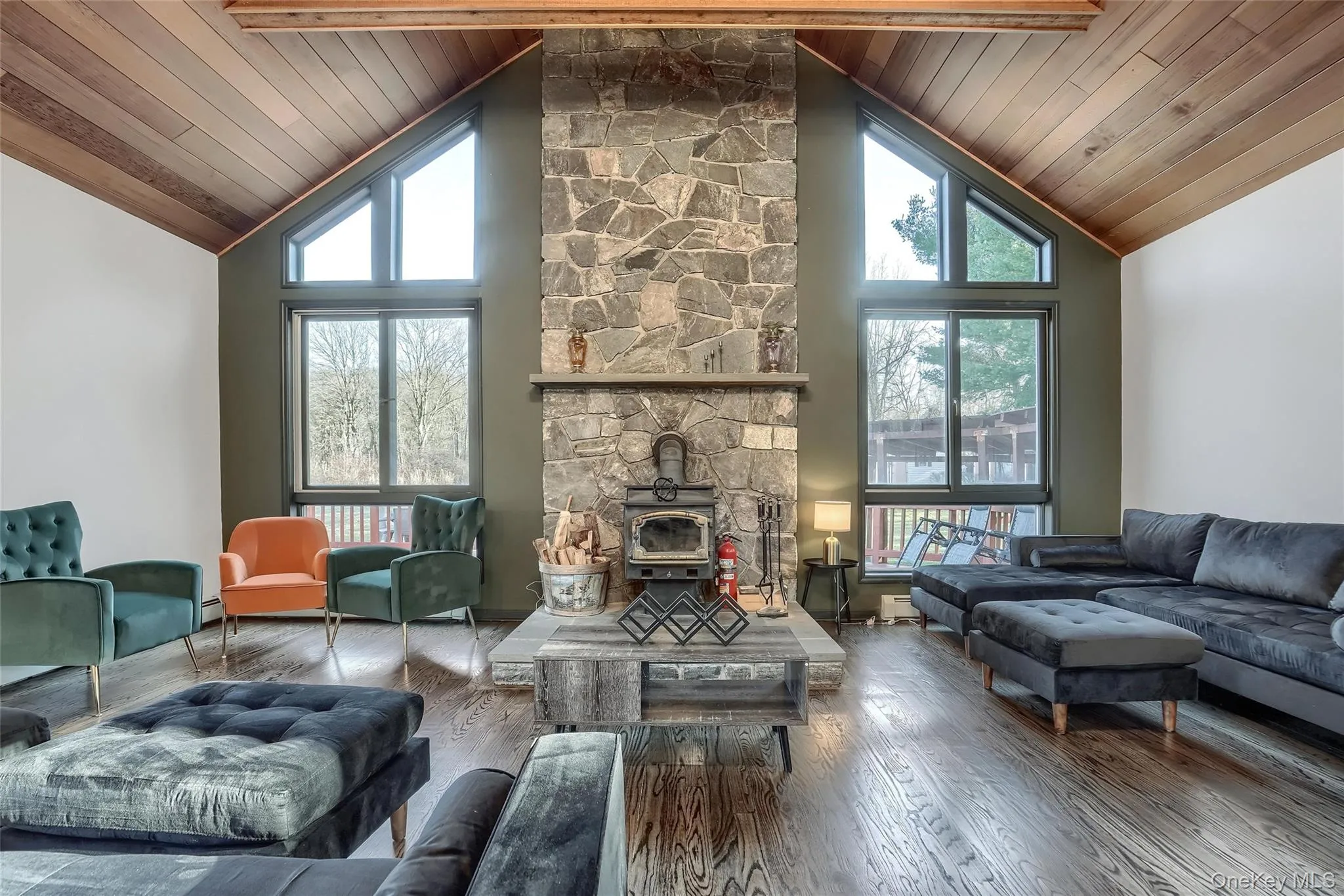 Vaulted wood-paneled ceiling with exposed beams, central stone-clad fireplace with wood-burning stove, and dark wood-finish flooring Vaulted wood-paneled ceiling with exposed beams, central stone-clad fireplace with wood-burning stove, and dark wood-finish flooring