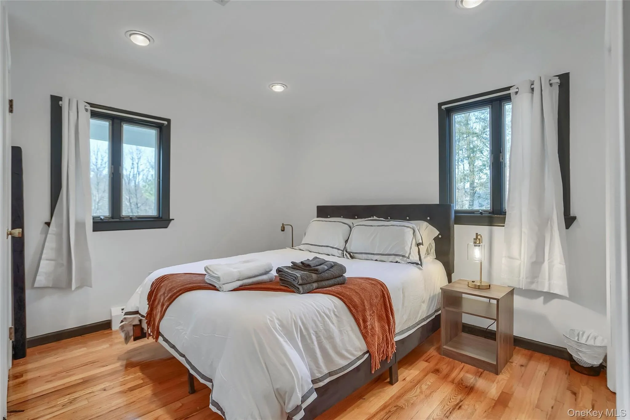 Bedroom featuring hardwood flooring, white walls, two windows with dark trim, and recessed ceiling lighting Bedroom featuring hardwood flooring, white walls, two windows with dark trim, and recessed ceiling lighting