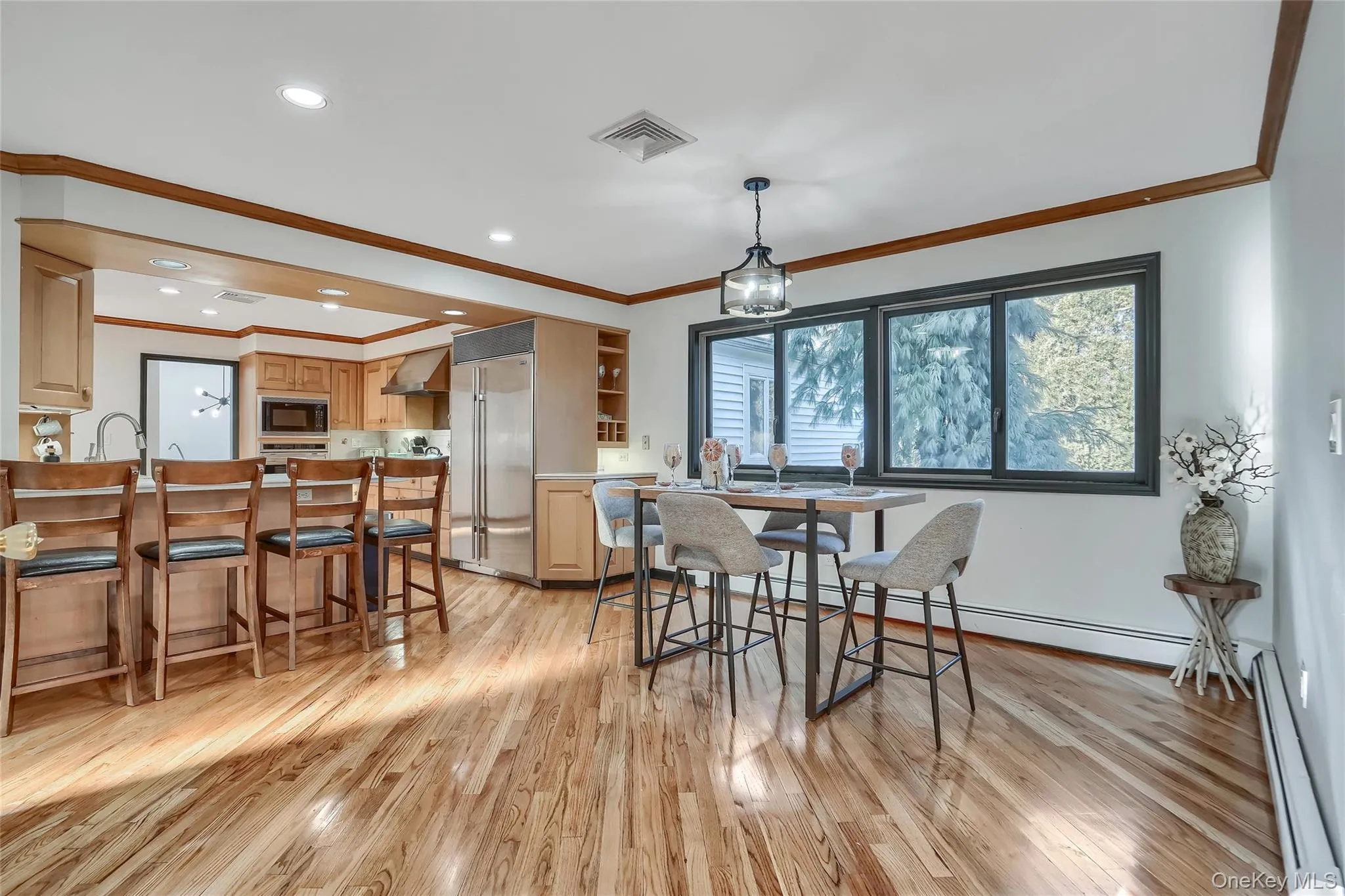 Open-concept kitchen and dining area featuring wood-finish flooring, recessed lighting, a breakfast bar with seating, stainless steel appliances, and large windows with dark trim Open-concept kitchen and dining area featuring wood-finish flooring, recessed lighting, a breakfast bar with seating, stainless steel appliances, and large windows with dark trim