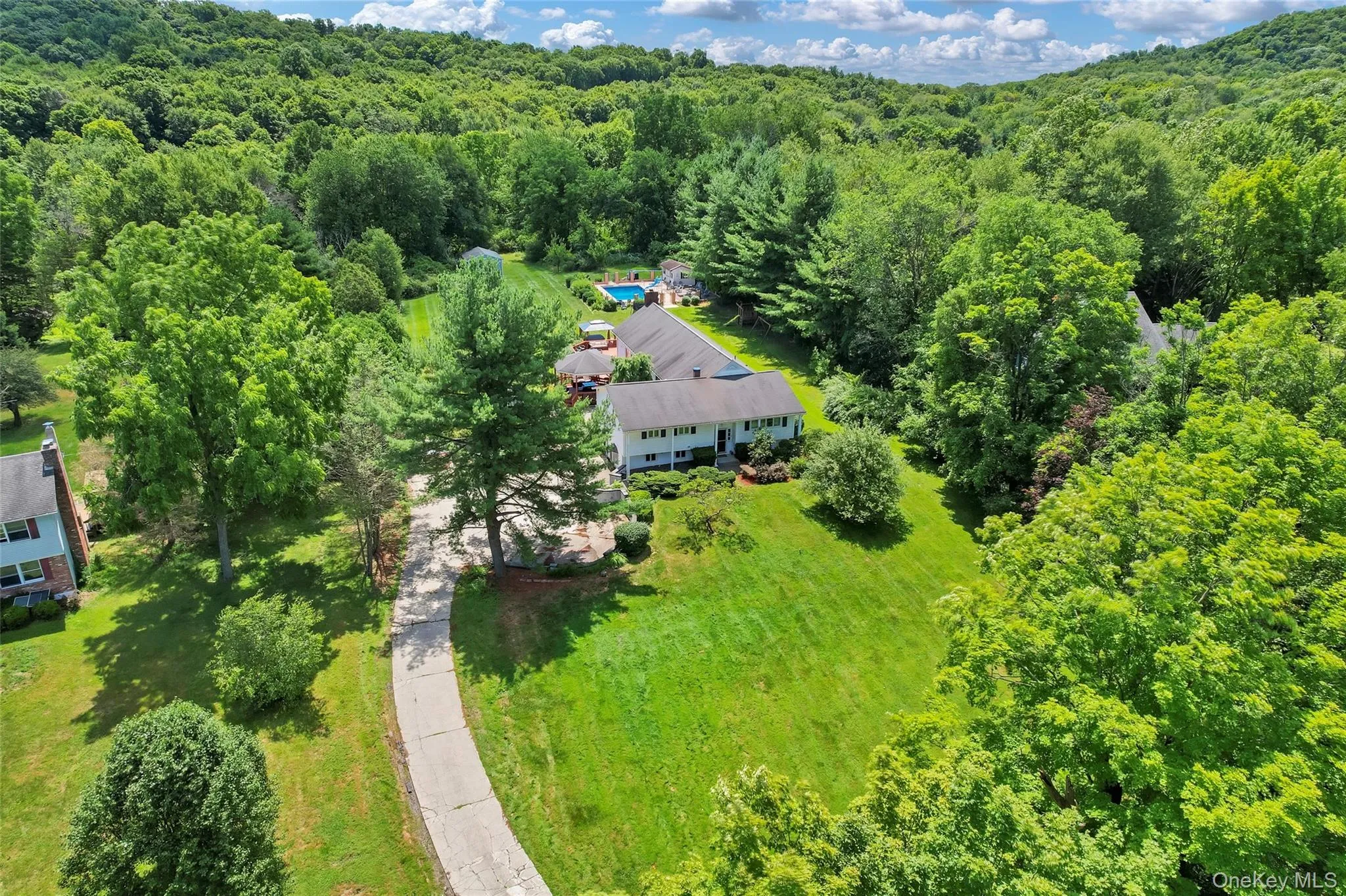 Expansive property featuring a single-story residence, a paved driveway, a swimming pool, and mature trees, all set against a backdrop of natural landscaping Expansive property featuring a single-story residence, a paved driveway, a swimming pool, and mature trees, all set against a backdrop of natural landscaping